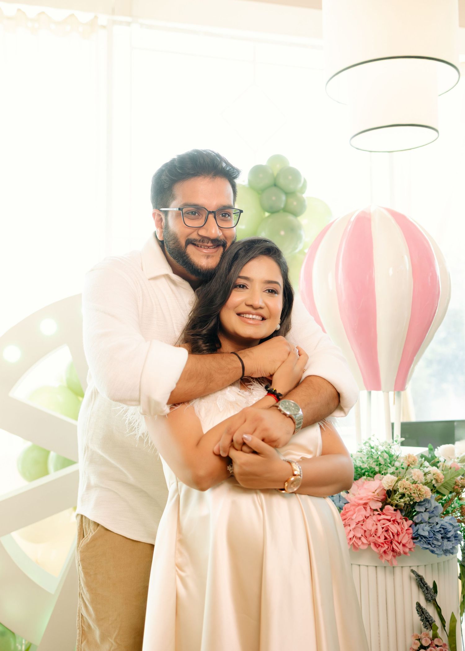 A couple embraces warmly at a baby shower, surrounded by pastel decorations, flowers, and a hot air balloon centerpiece.