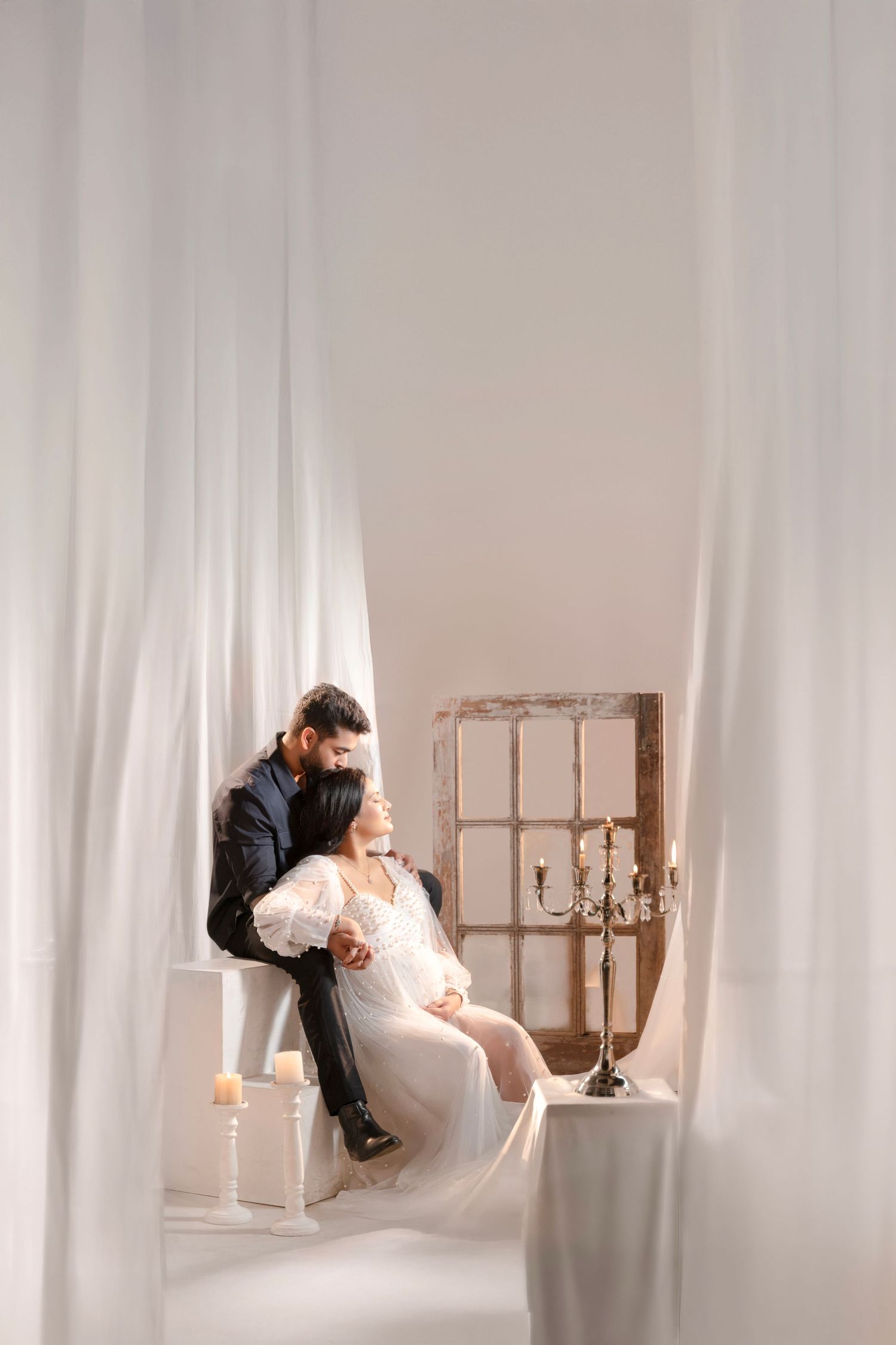 Elegant couple in white studio setting, groom in dark suit embracing bride in flowing white gown near vintage window decor.
