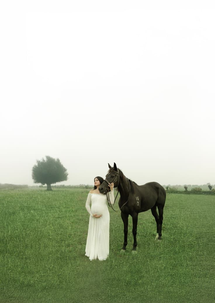 Pregnant woman in white gown stands beside a dark horse in a misty green field with a lone tree.