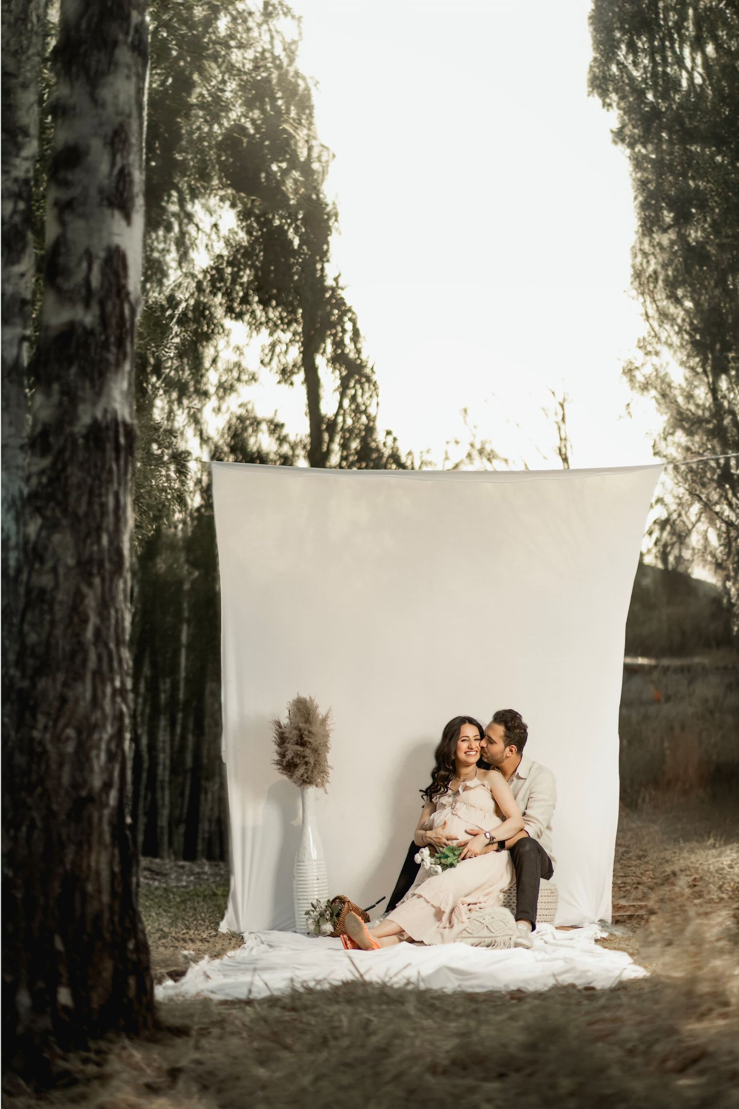 A romantic outdoor photo session featuring a couple seated on white fabric against a minimalist backdrop in a wooded setting.