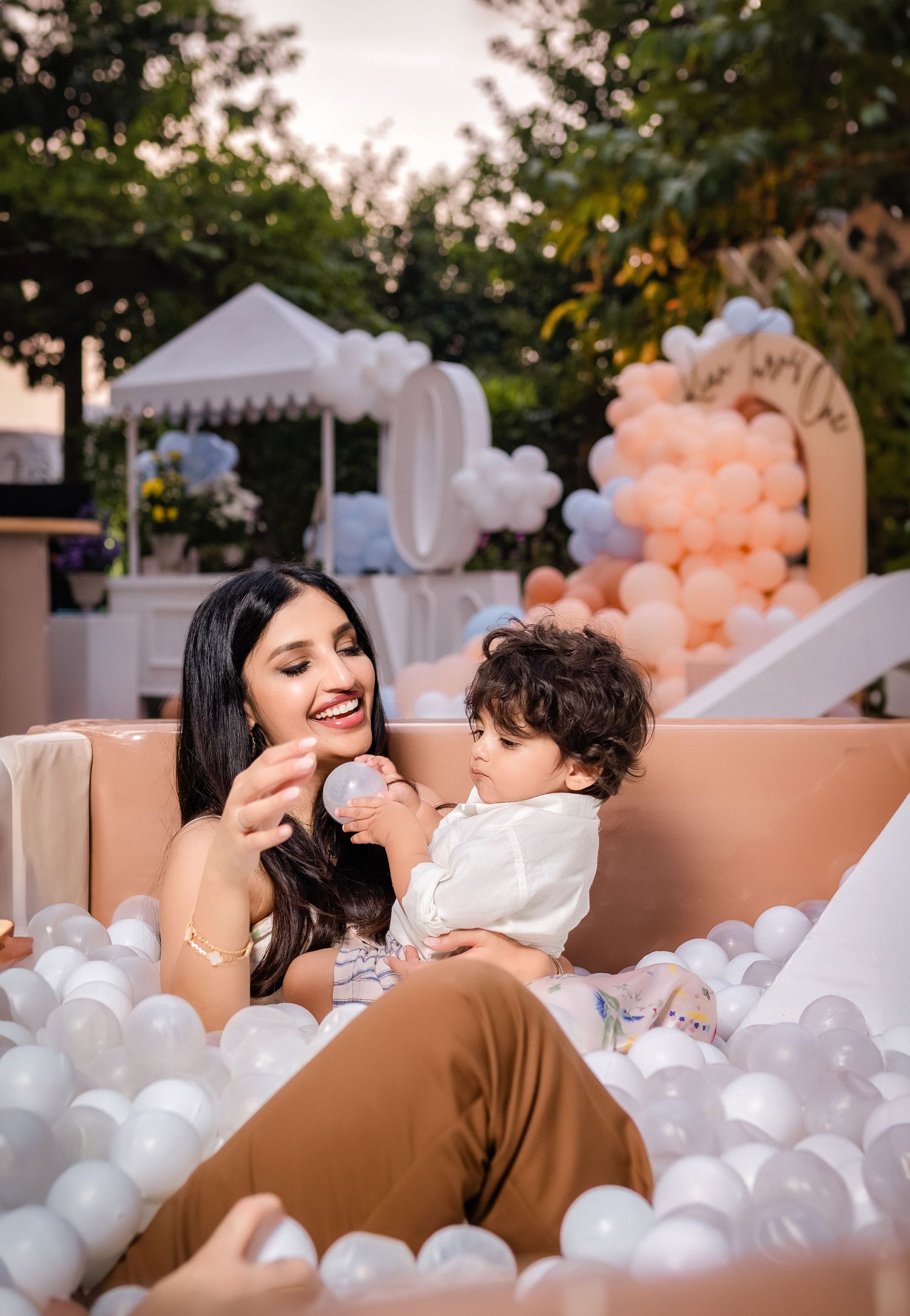 A parent and child sitting together in a ball pit filled with white balloons at an outdoor event with peach decor.