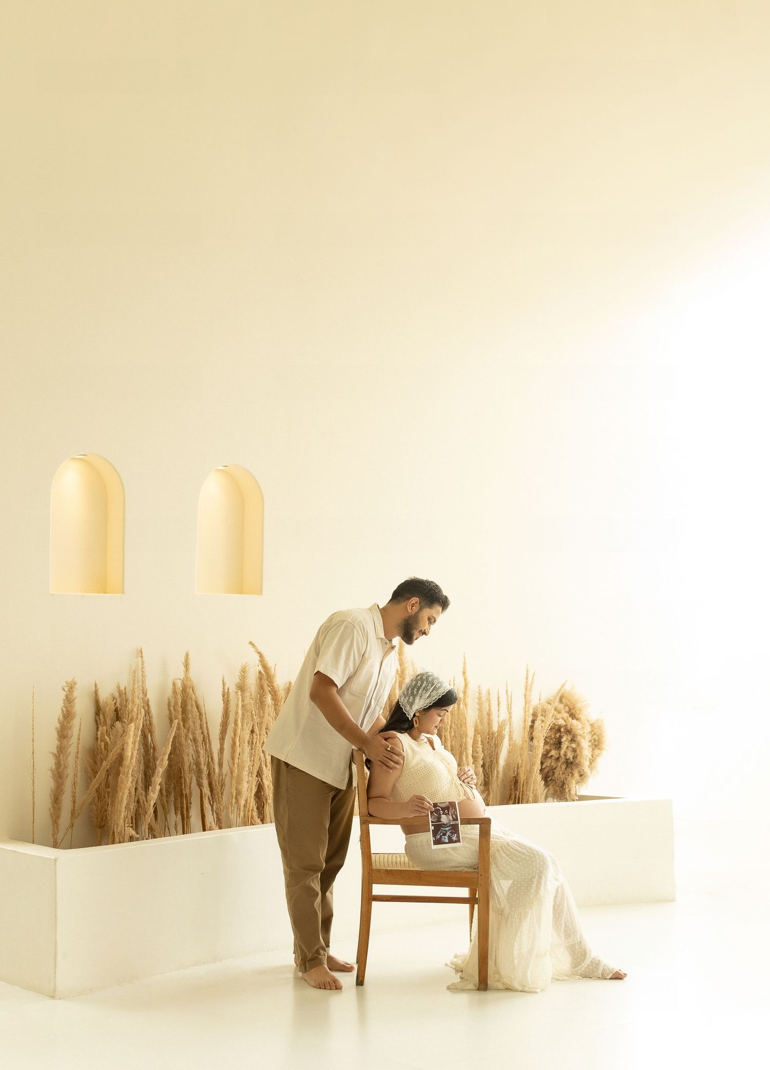 A person in beige clothing examines a display of dried wheat stalks arranged against a white wall in a minimalist setting.