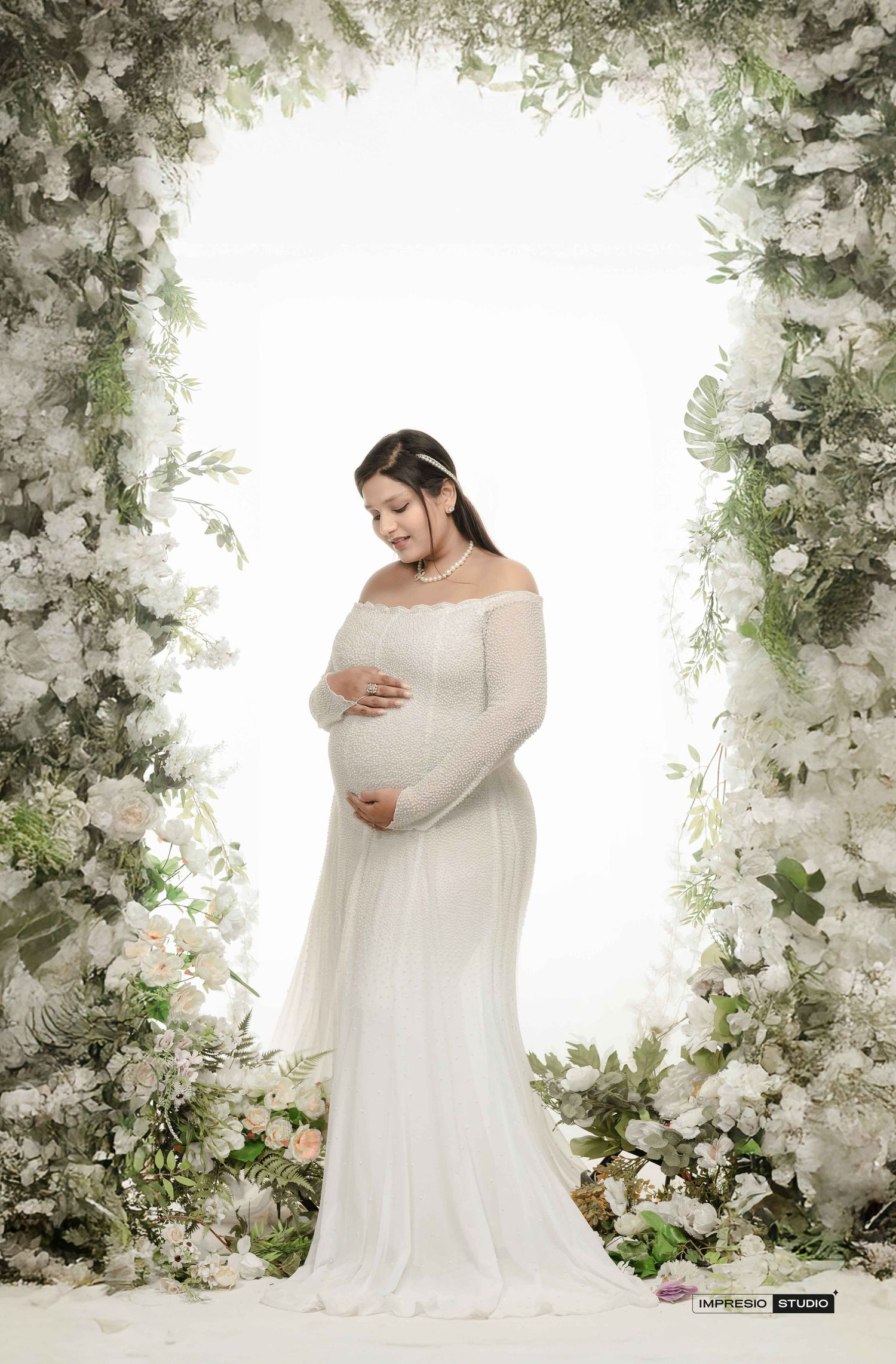 Pregnant woman in white off-shoulder gown poses gracefully under a floral arch with white blooms and greenery.