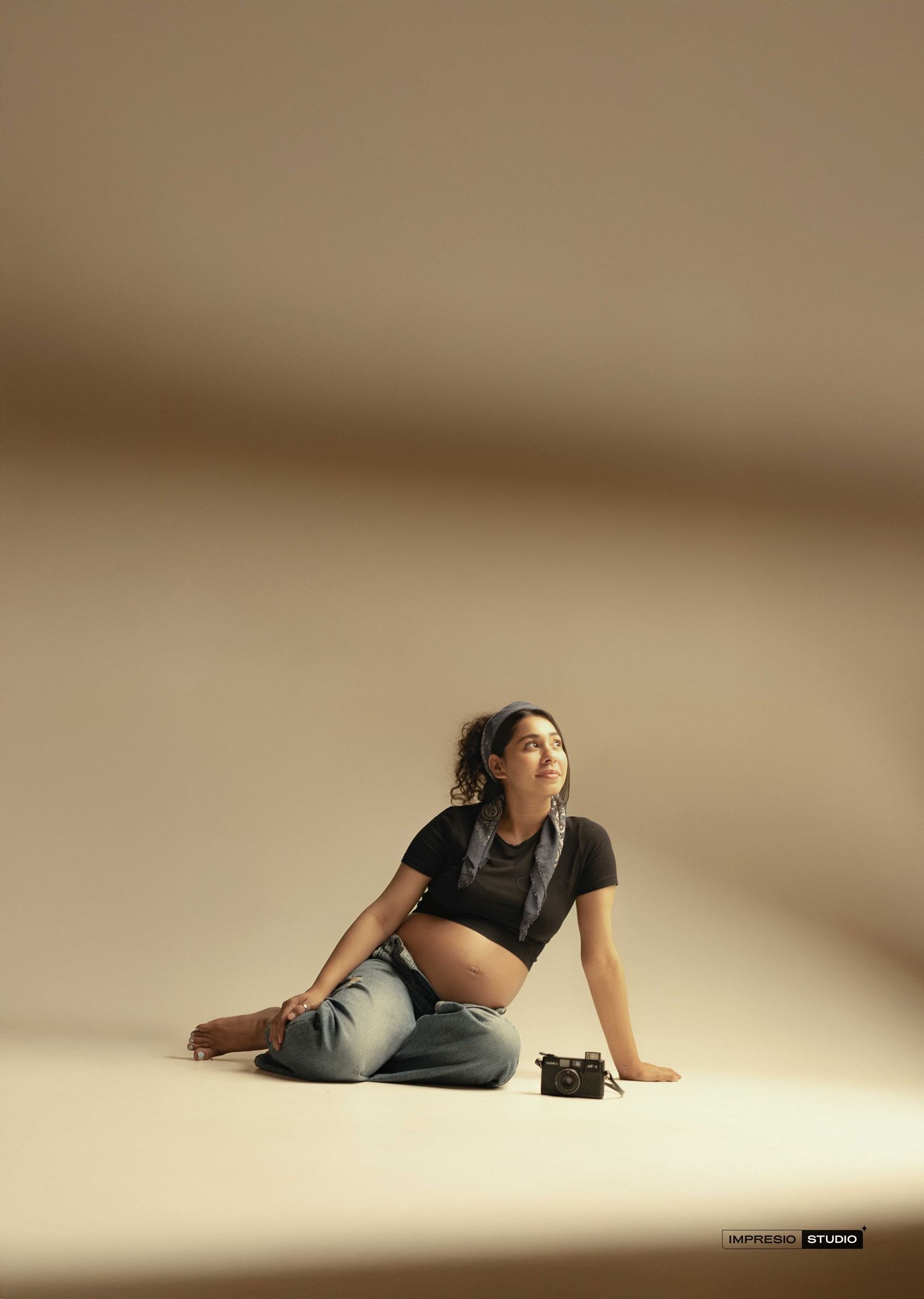 Pregnant woman in black crop top and jeans sitting on studio floor with vintage camera beside her.