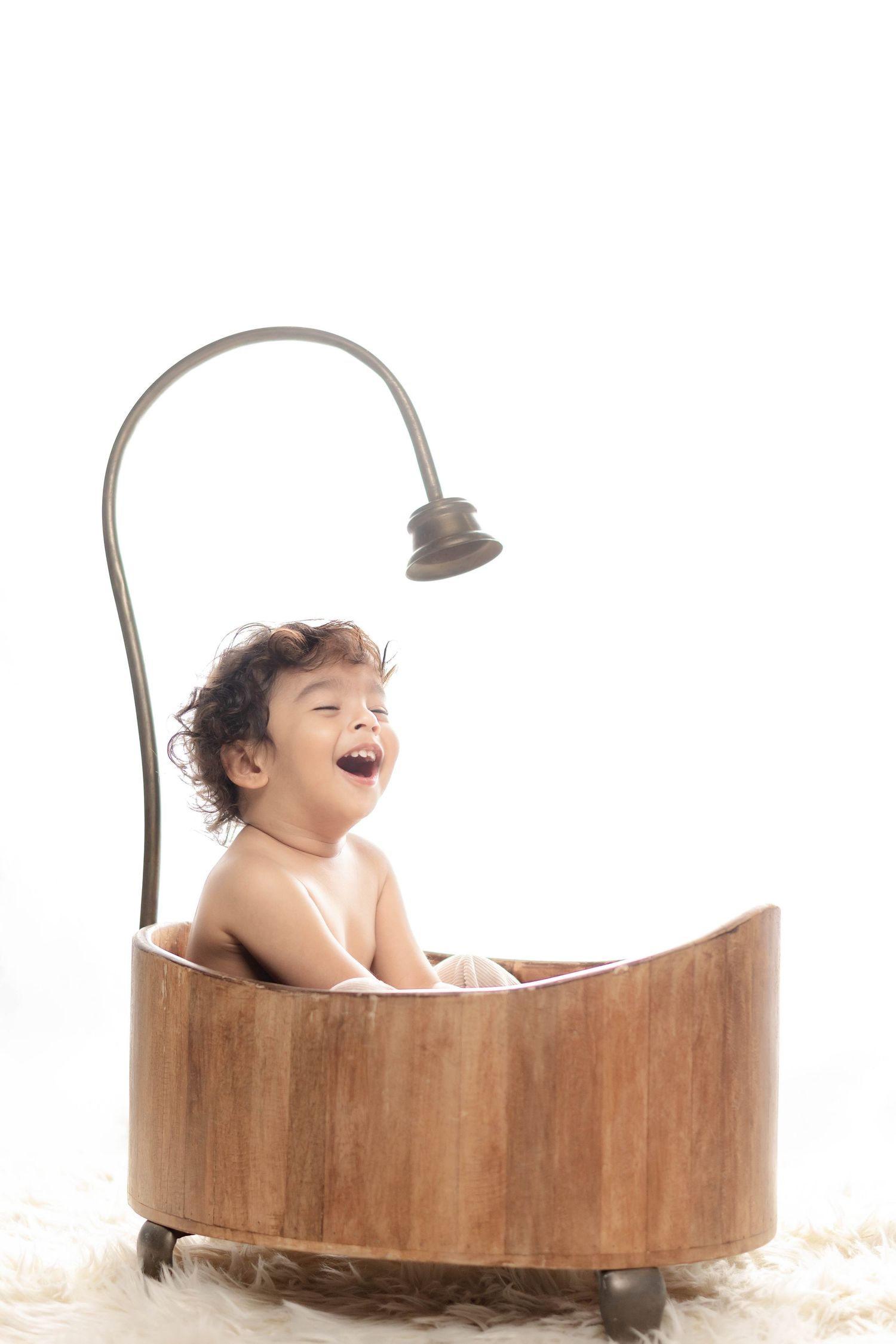 Laughing baby sitting in a rustic wooden bathtub with a vintage shower head on a white background.