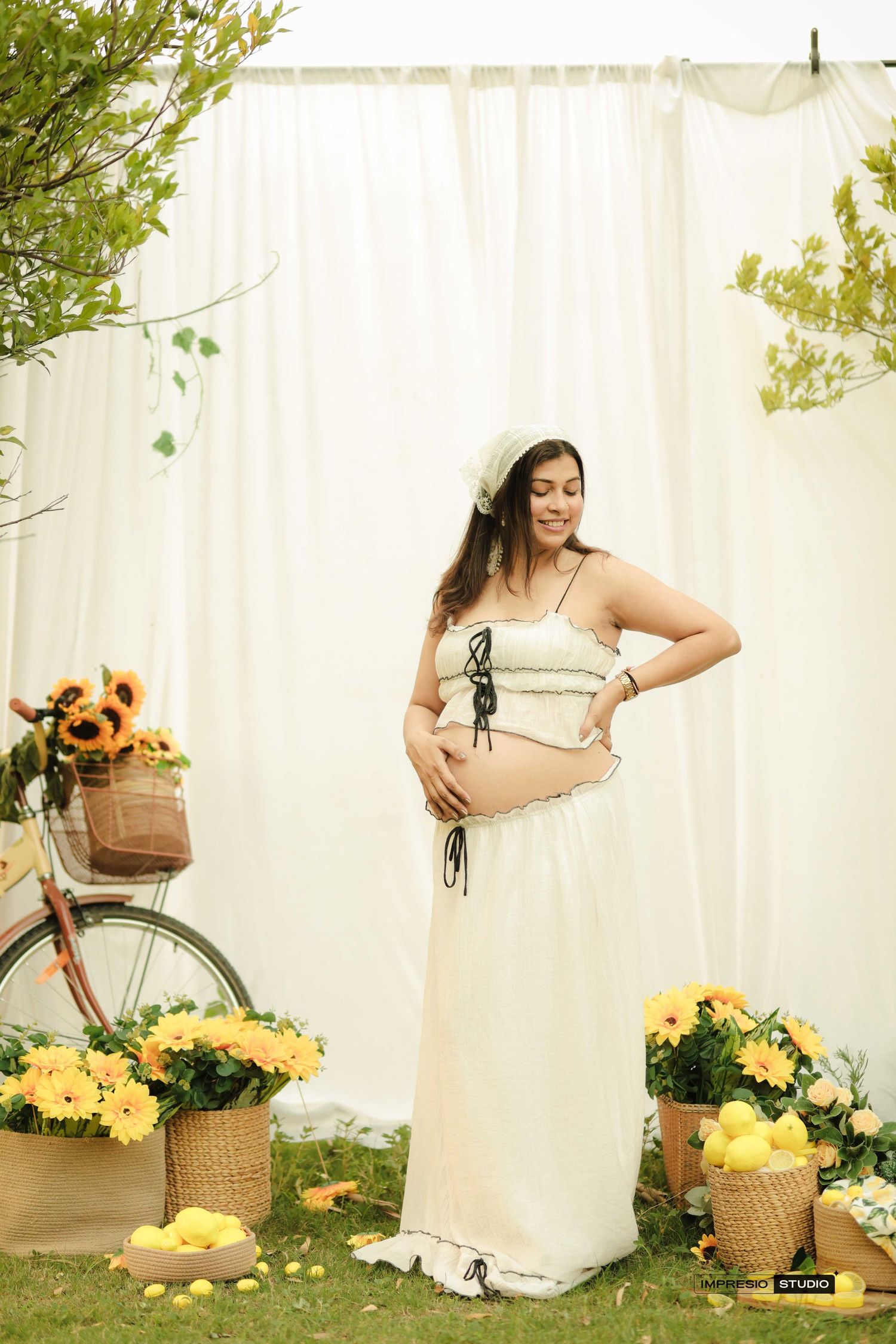 Pregnant woman in cream crop top and skirt cradling baby bump surrounded by yellow flowers outdoors.