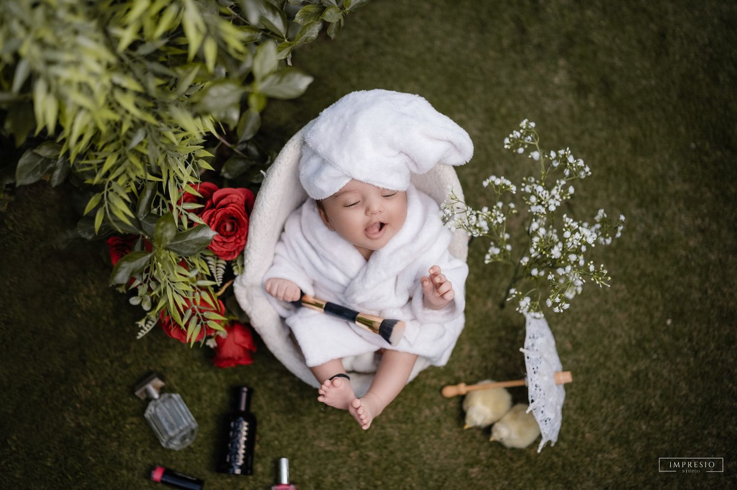 Newborn baby in white robe and bunny ears sleeping in grass surrounded by red flowers and small props.