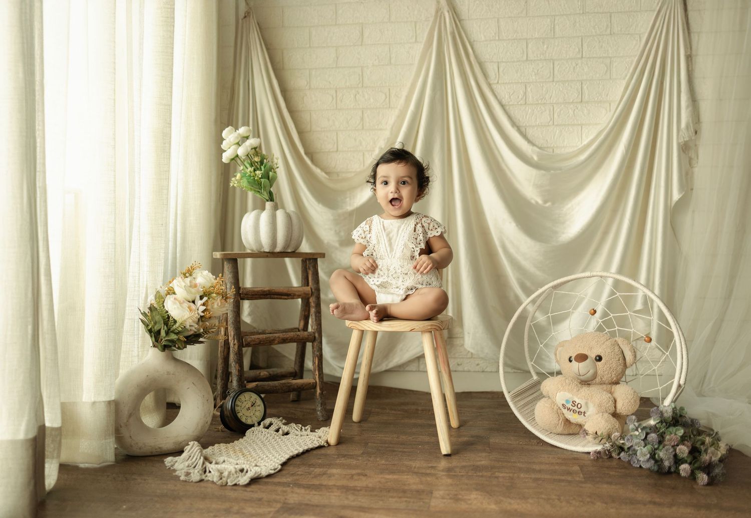 A cheerful baby sits on a wooden chair surrounded by soft decor, flowers, and a teddy bear in a bright studio setting.
