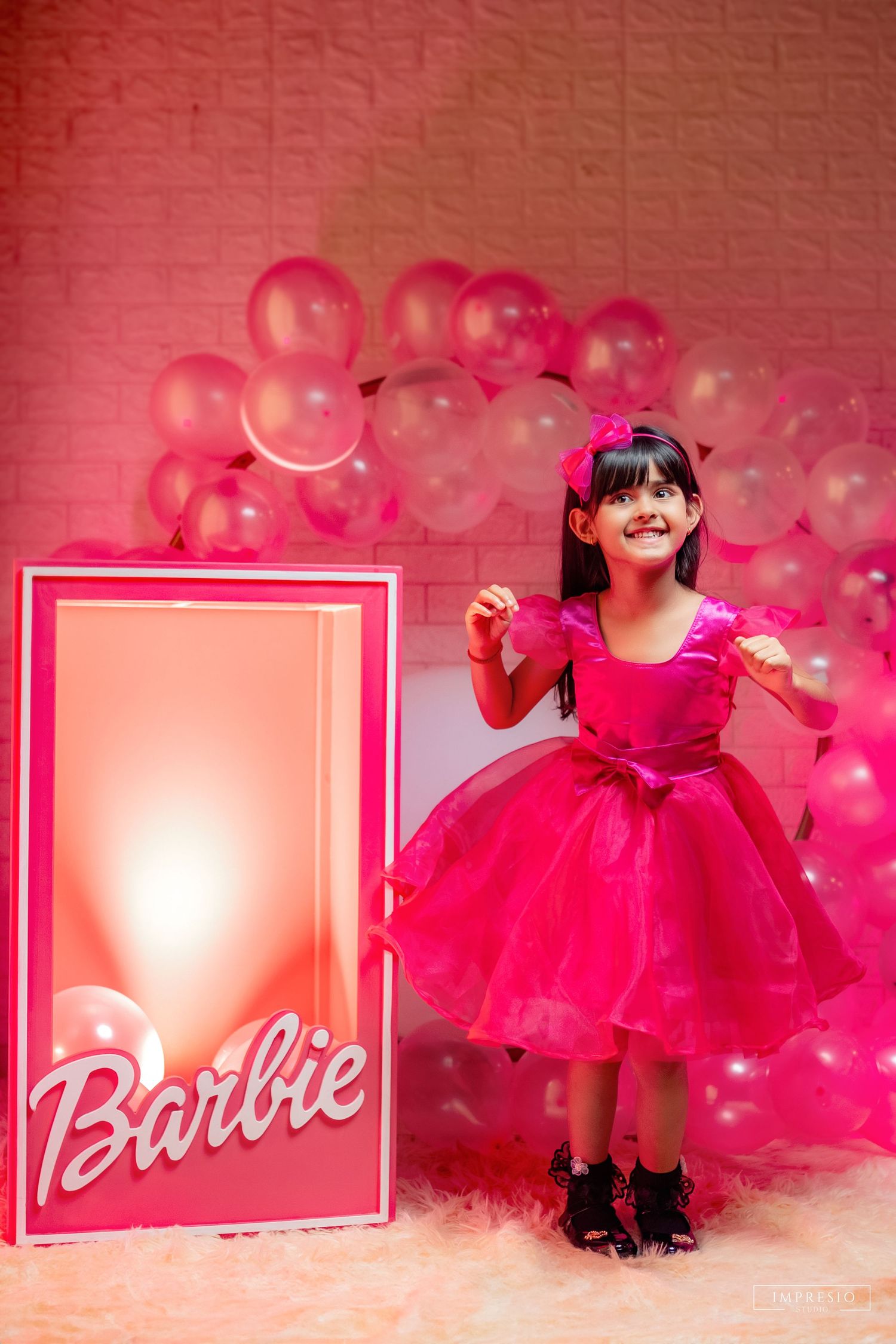 A young girl in a hot pink Barbie dress poses beside a Barbie box prop surrounded by pink balloons at a themed birthday party.