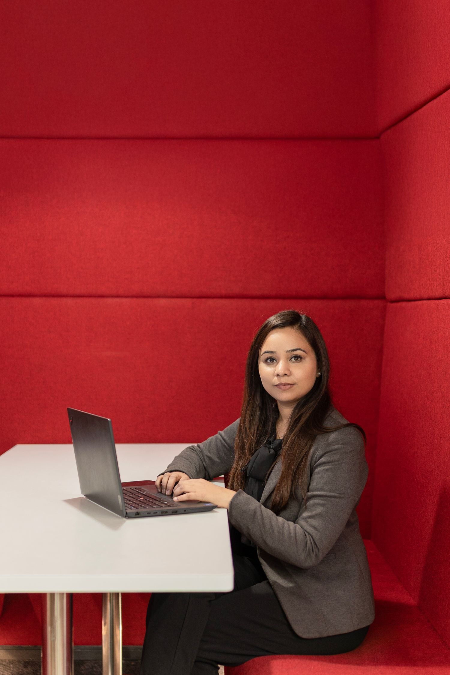 Professional in business attire works on laptop computer while seated at white desk against vibrant red wall.
