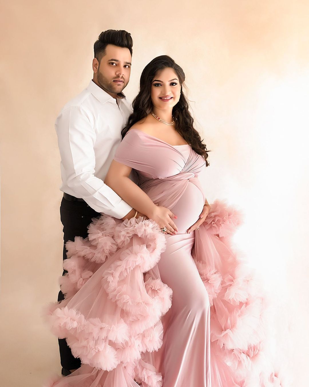 Expectant couple poses in formal attire with flowing pink tulle gown against neutral background.