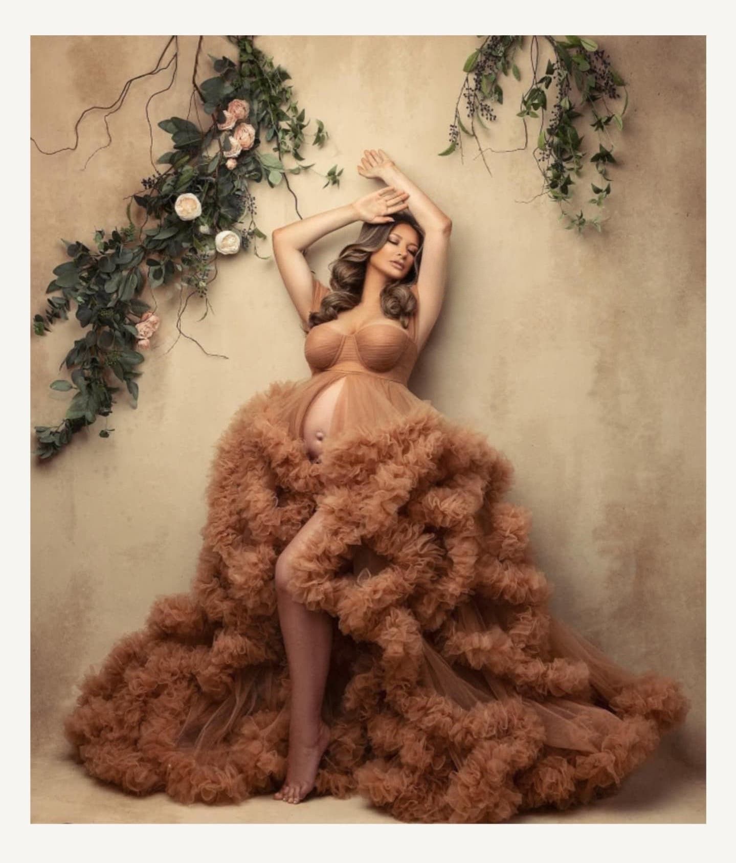 Artistic maternity photo with floral decor and a flowing copper-colored tulle dress against a neutral backdrop.