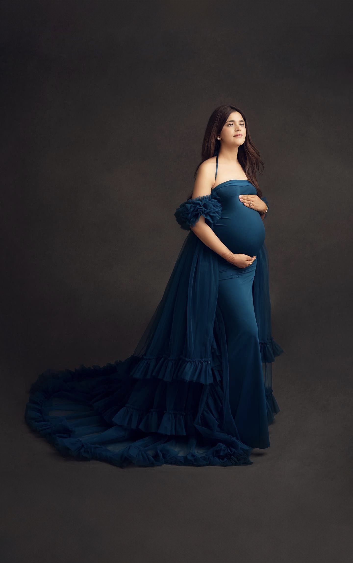 Dramatic maternity portrait in a flowing navy blue off-shoulder gown with ruffle details against dark background.