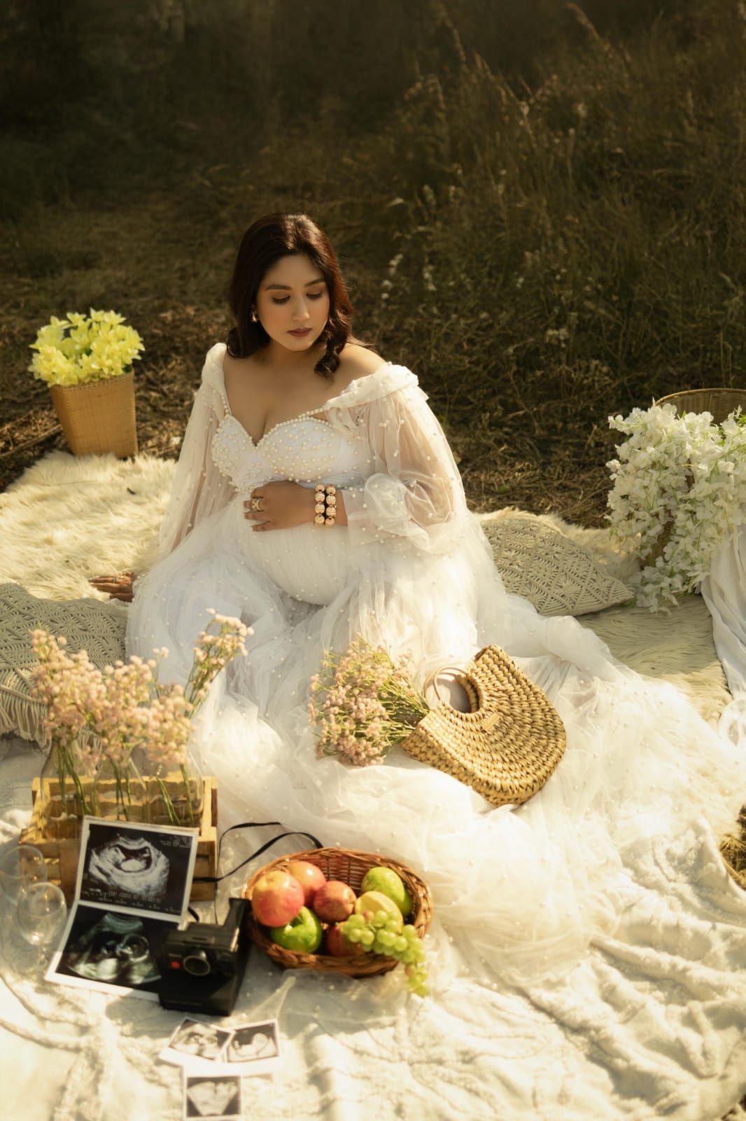 Maternity picnic scene with white dress, fruit basket, and ultrasound photos on blanket.