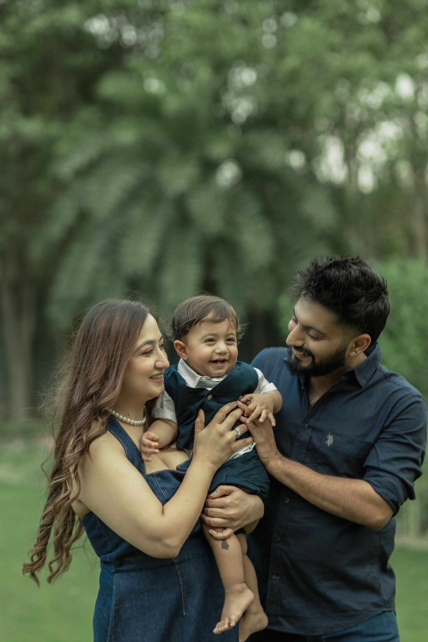 Happy Indian family with parents holding laughing baby boy outdoors in lush green park setting.