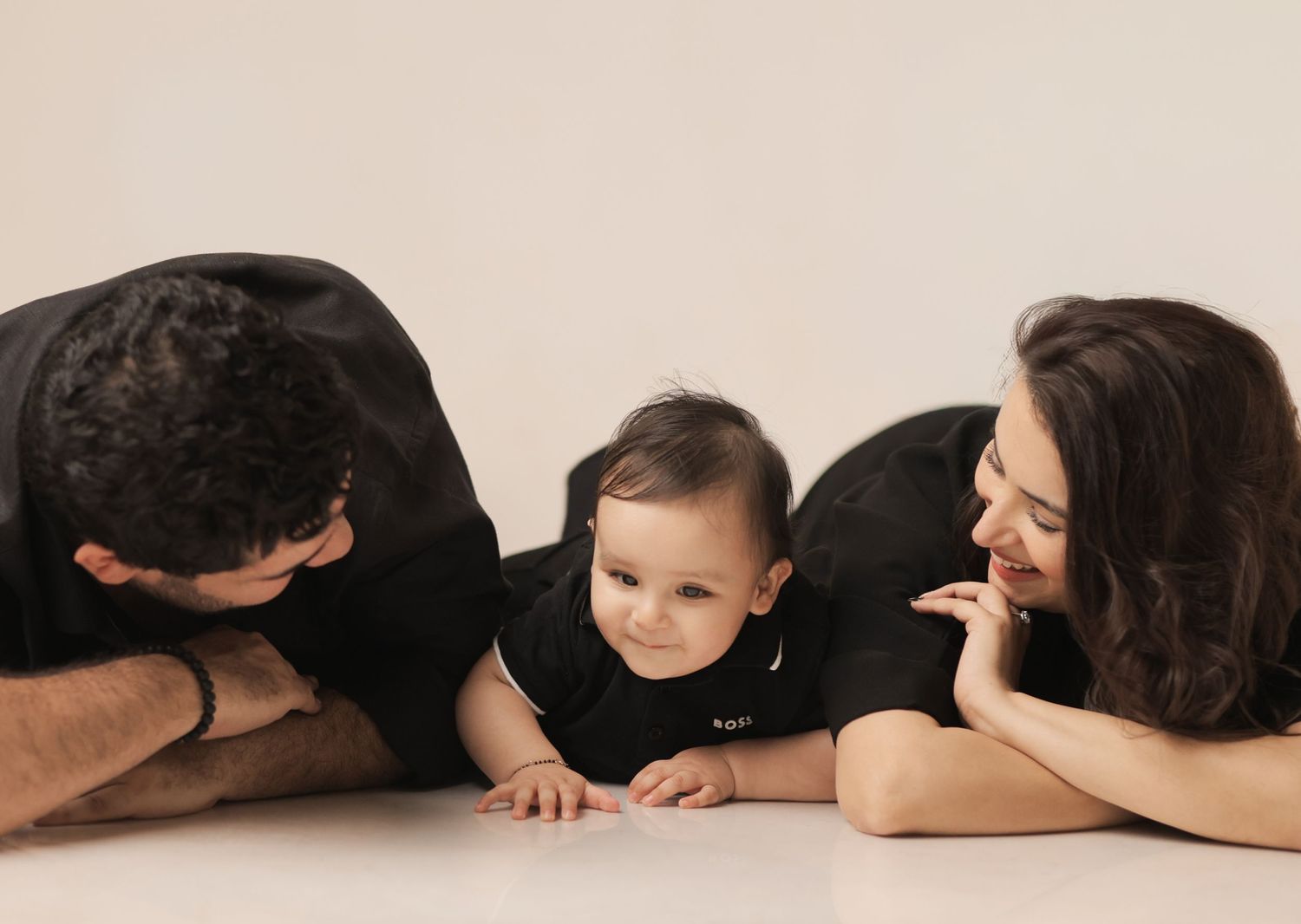 Family photo of parents in black outfits lying on floor with smiling baby in black onesie on white background.