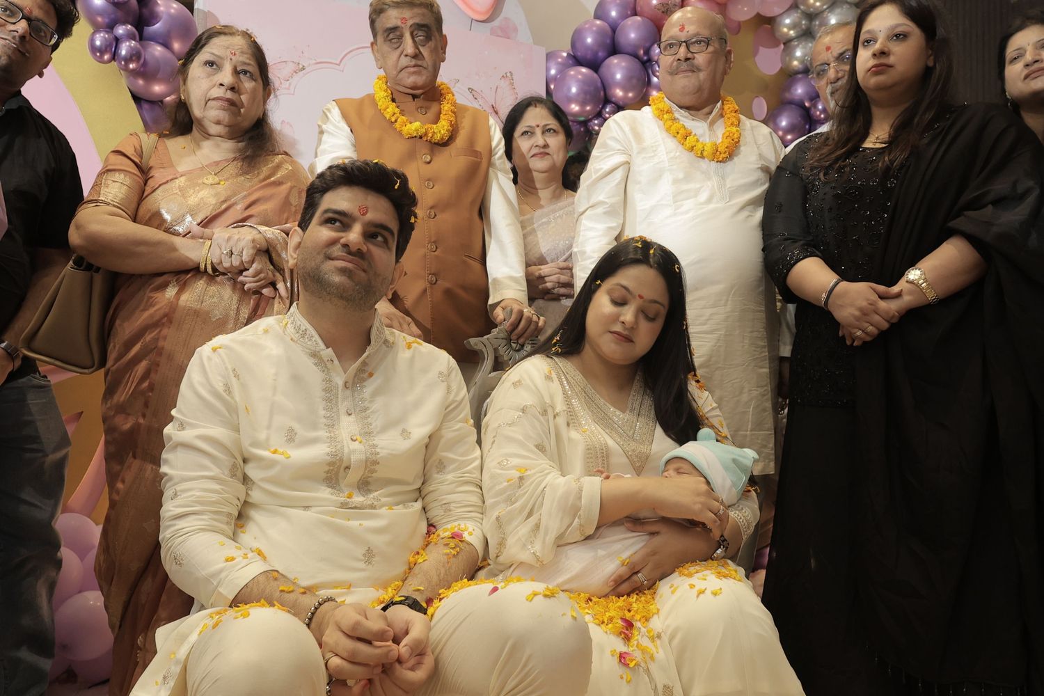 A smiling couple in cream traditional attire sits with a newborn while family stands behind them near purple and white balloons.