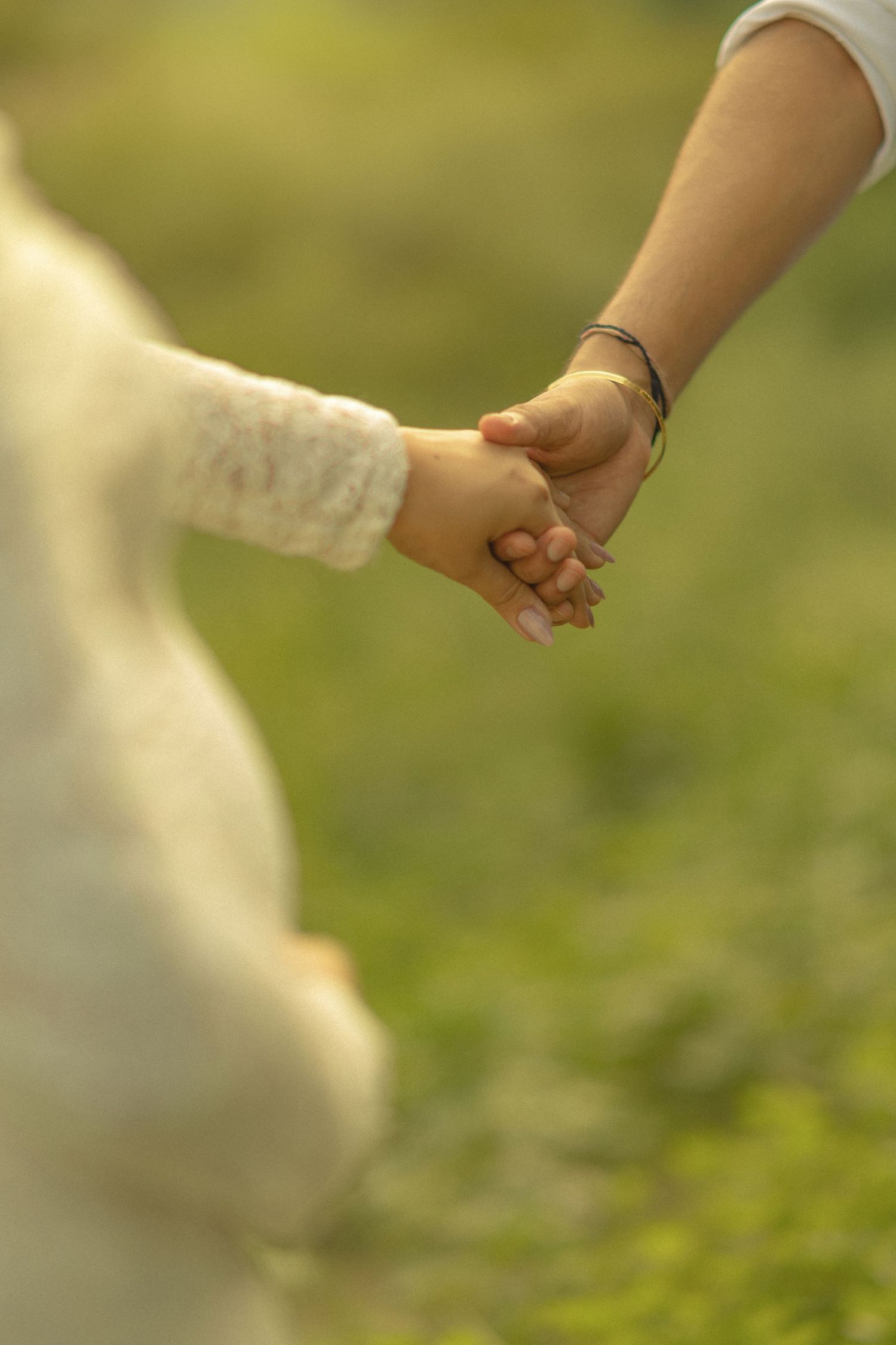 Adult and child holding hands outdoors in a green field, wearing bracelets, in warm natural light.