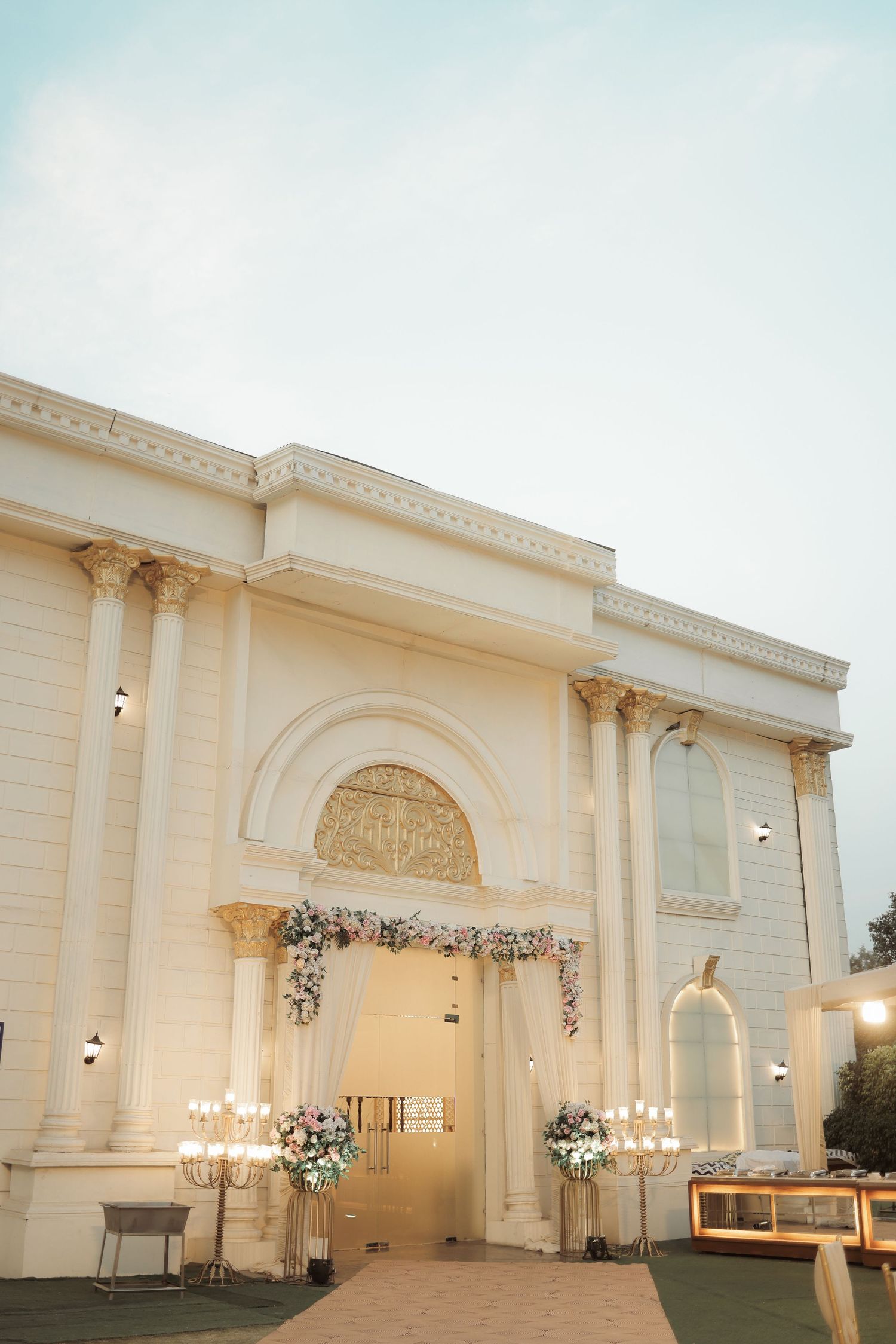 Elegant white wedding venue exterior with floral decorations and chandeliers at dusk.
