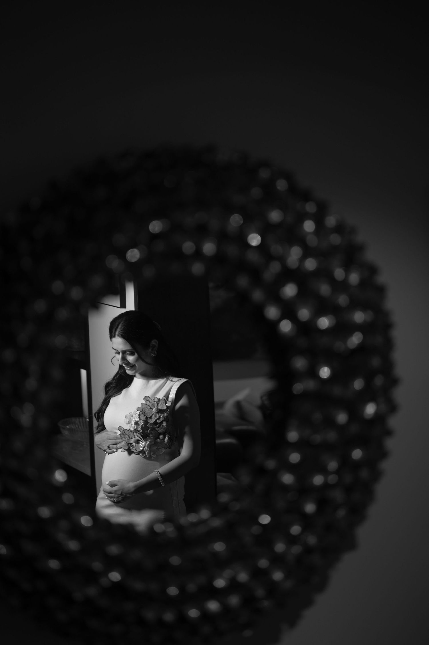 Black and white mirror reflection of a pregnant woman holding flowers, framed by a decorative wreath.