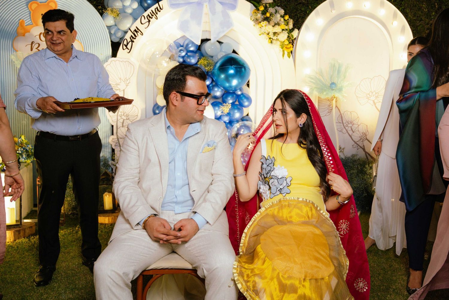 Couple in traditional attire at a baby shower celebration with blue balloon decorations.