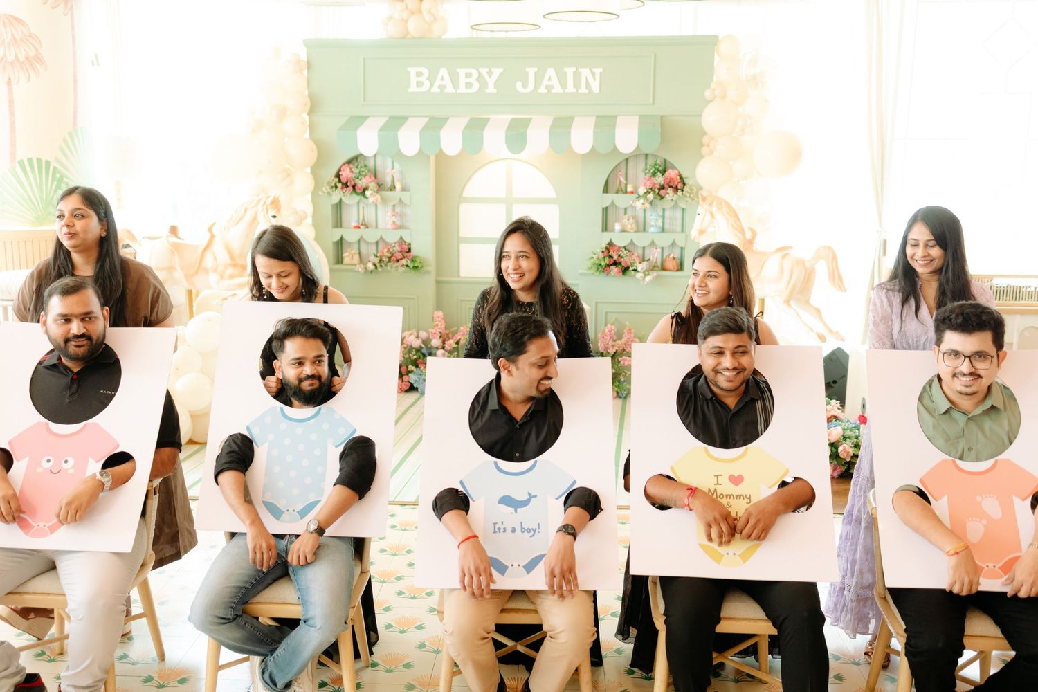 Group of friends posing with baby bib cutouts at a Baby Jain shower celebration in a decorated venue.