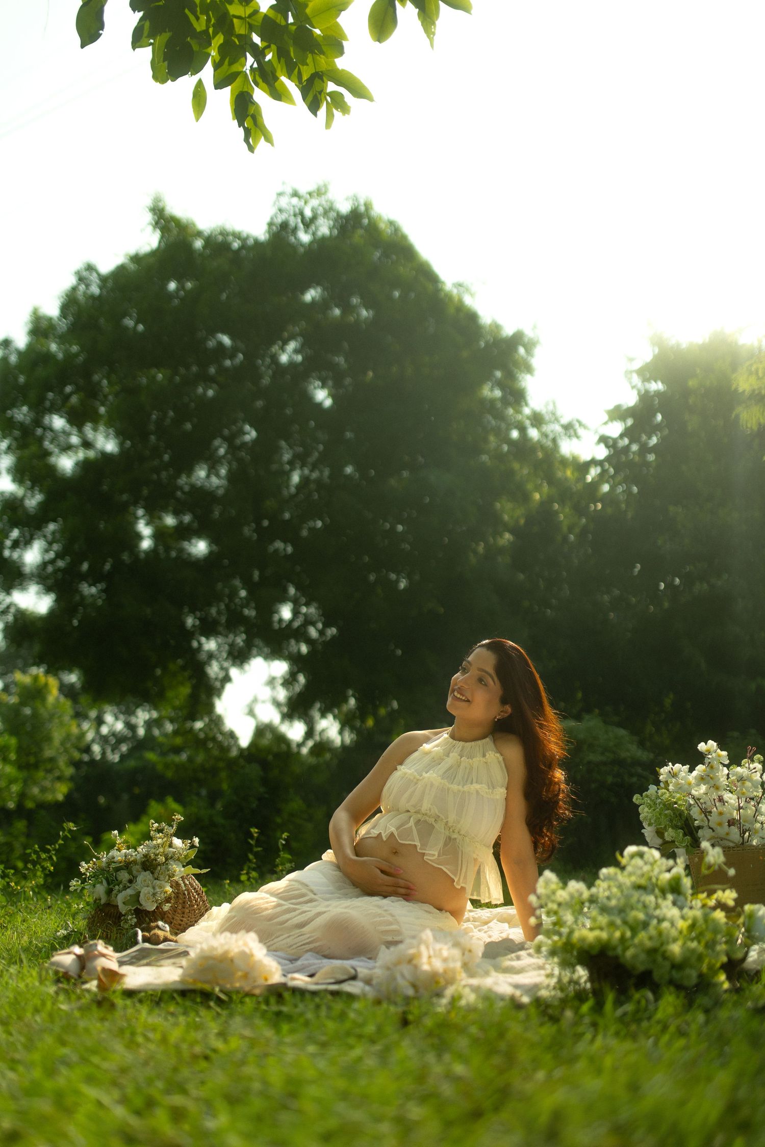 Pregnant woman in white dress sitting on grass in sunlit park, smiling and cradling her baby bump surrounded by flowers.