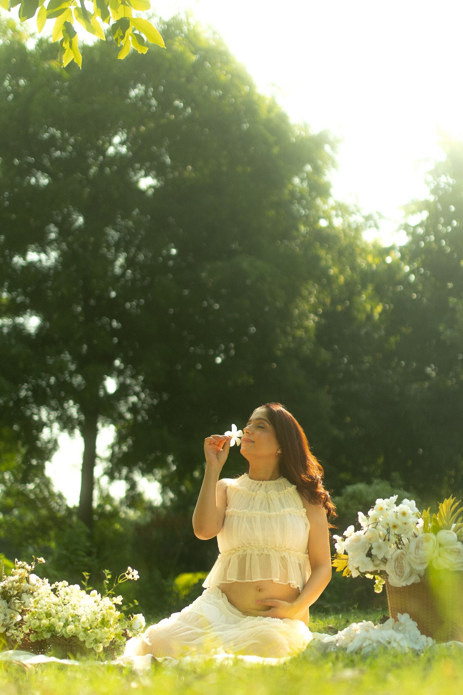 Pregnant woman in white dress sitting on grass, smelling a white flower in a sunlit park.