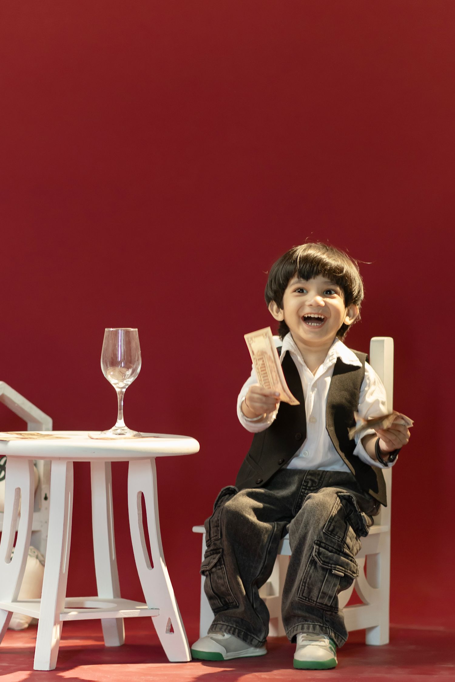 Happy young boy in suit sitting on white chair holding snacks beside a small table with wine glass on red background.