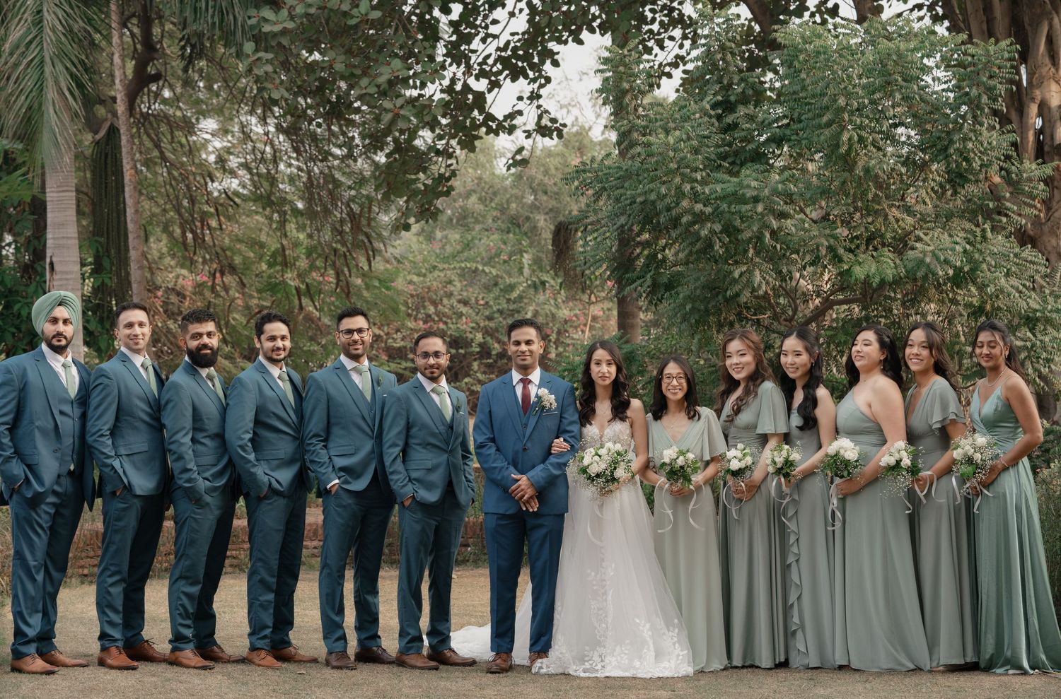 Wedding party in sage green bridesmaid dresses and gray groomsmen suits pose together on a tree-lined path.