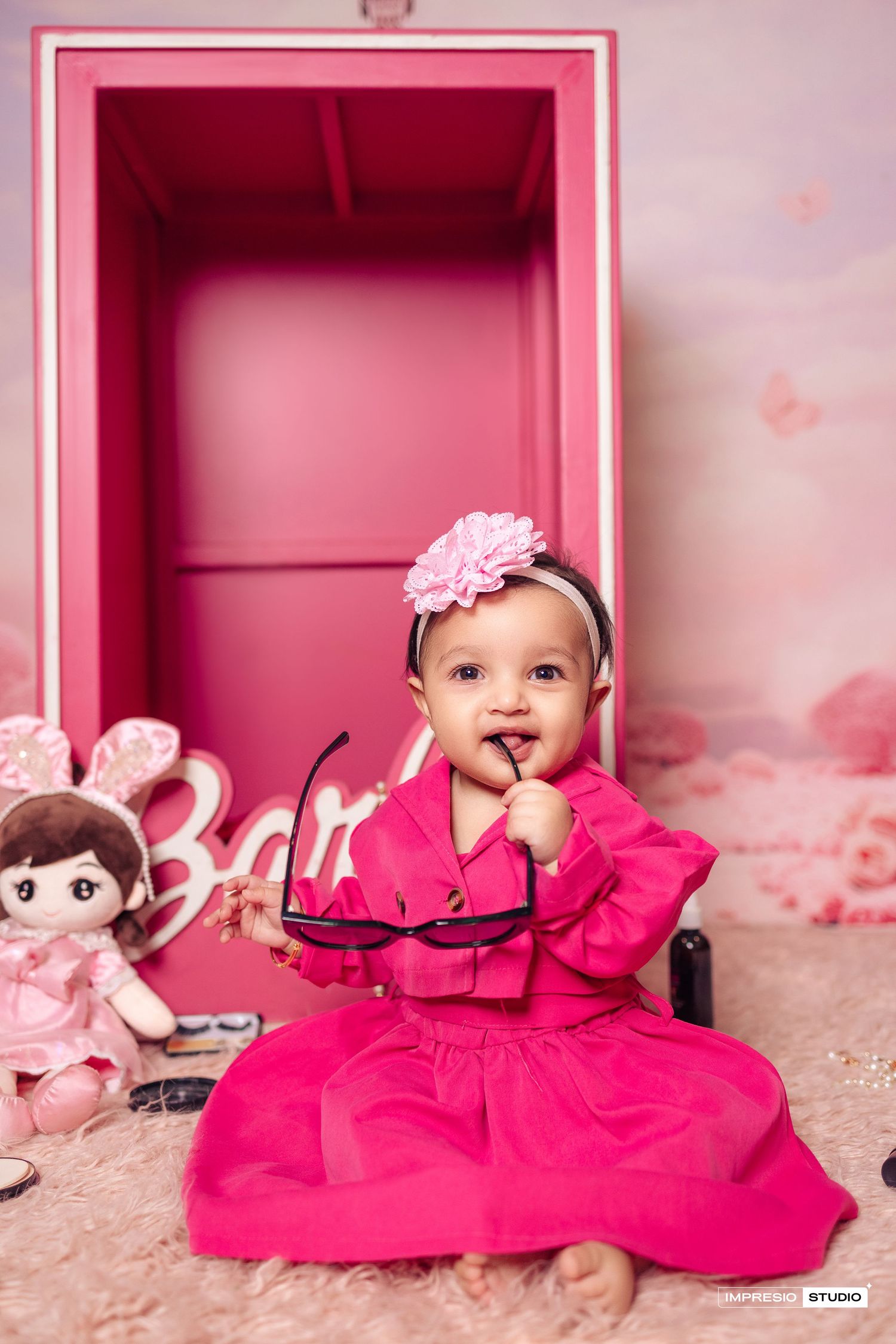 A baby in a bright pink dress sits on a plush carpet with a doll and pink decor in the background.