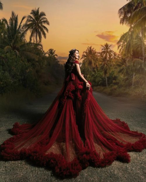 A figure in flowing burgundy gown poses dramatically against a tropical sunset with palm trees silhouetted in the background.