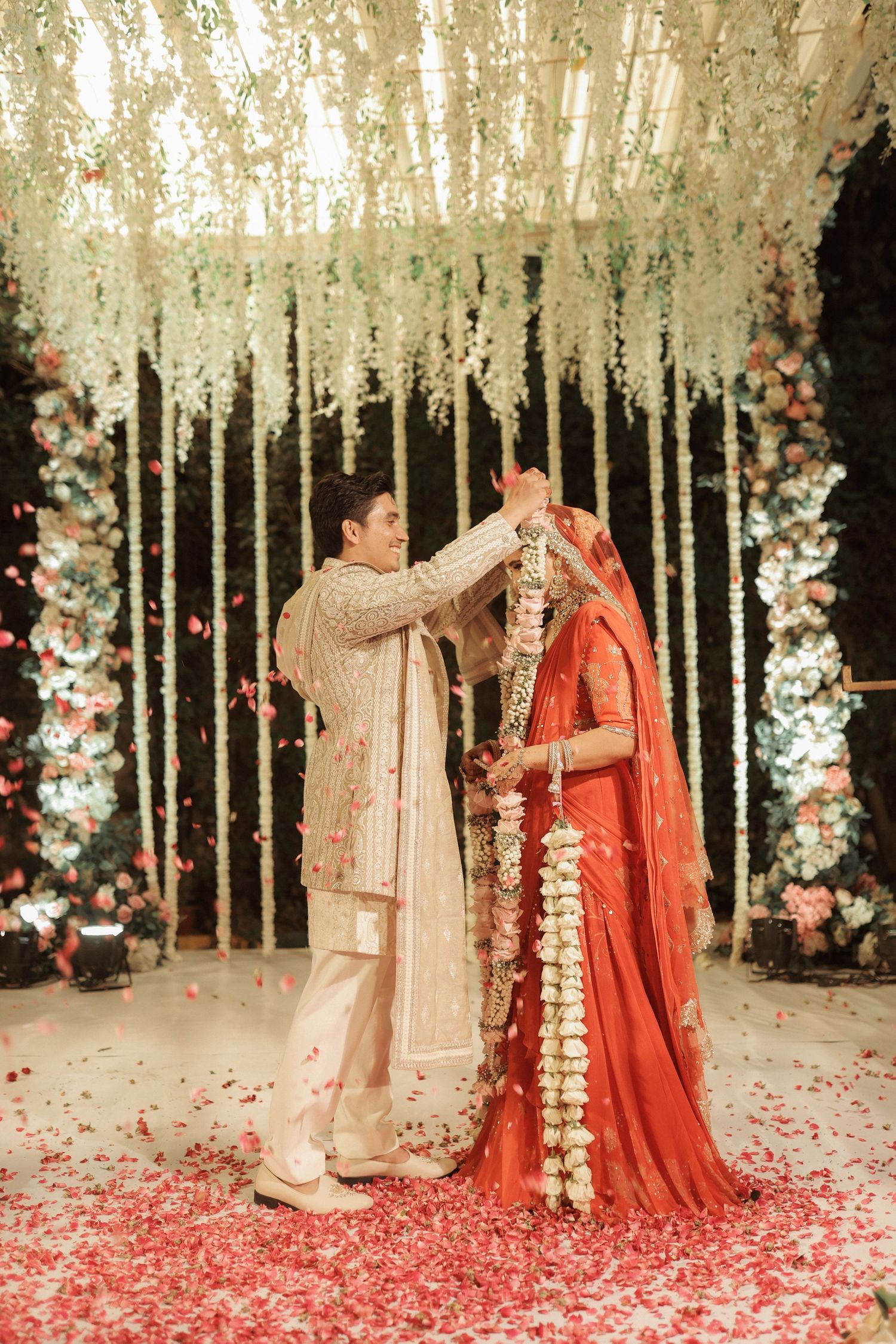 Indian couple exchanging garlands during wedding ceremony with floral backdrop and rose petals.
