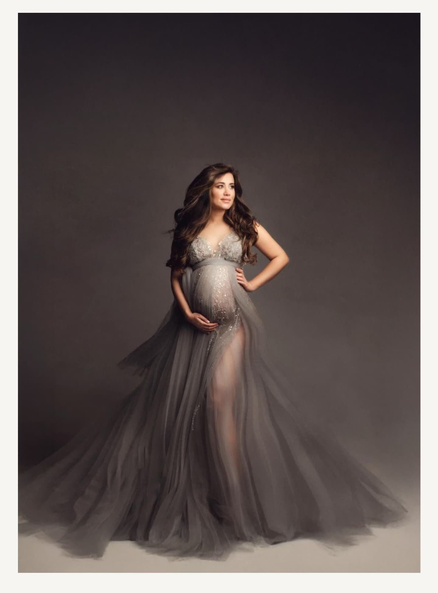 Glamorous maternity portrait in flowing gray tulle gown with dramatic lighting.