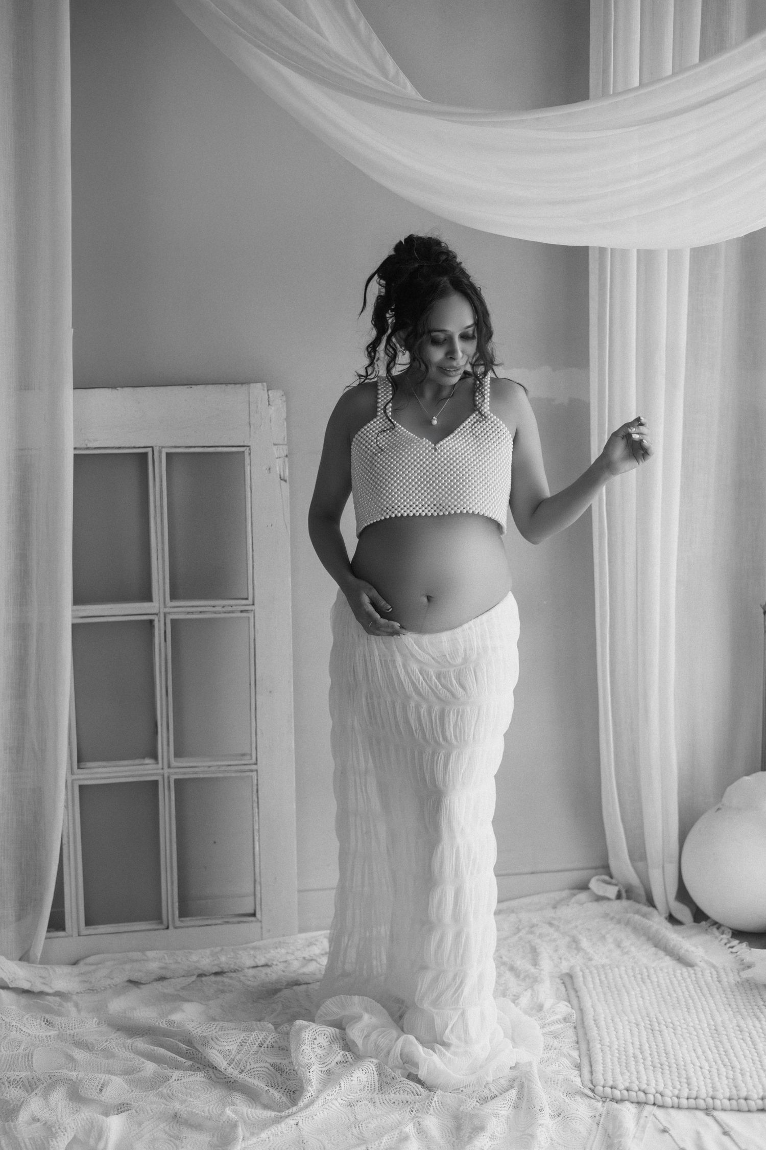 A series of black and white maternity photographs showing a pregnant woman in a white dress against light curtains.