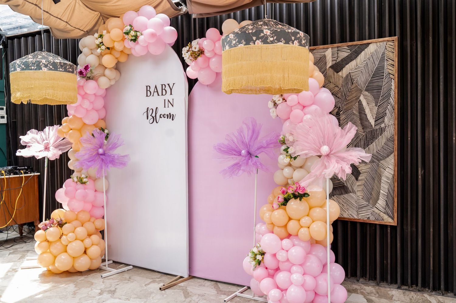 Baby shower balloon arch with pink, purple, and orange balloons, large paper flowers, and a 'Baby in Bloom' backdrop.