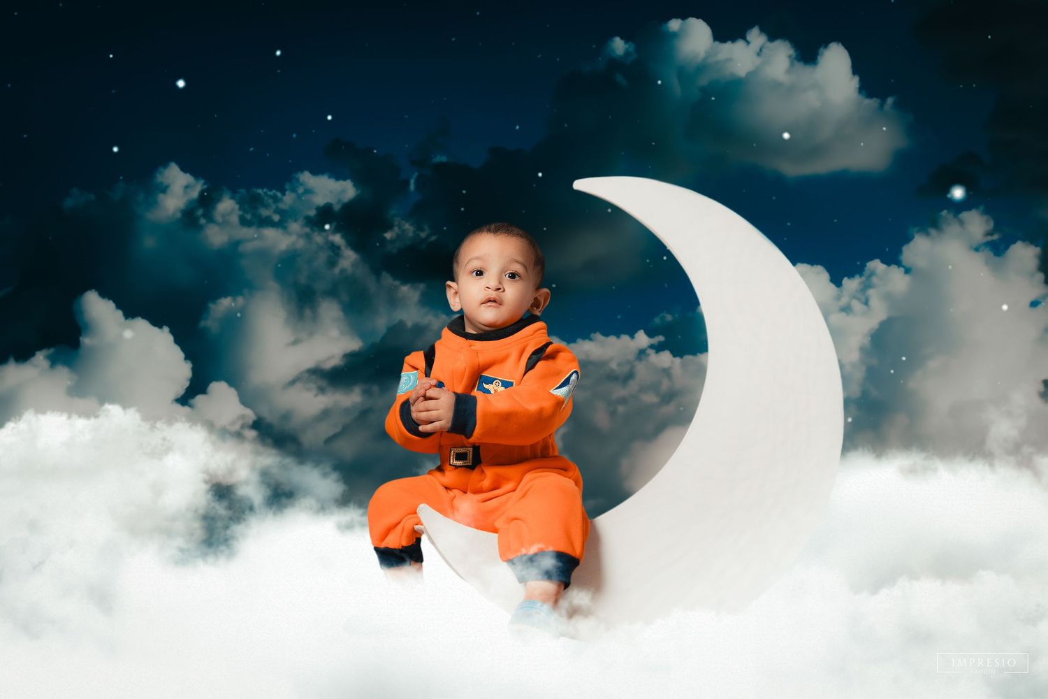 A toddler in an orange astronaut suit sits on a crescent moon prop surrounded by clouds in a dreamy night sky.
