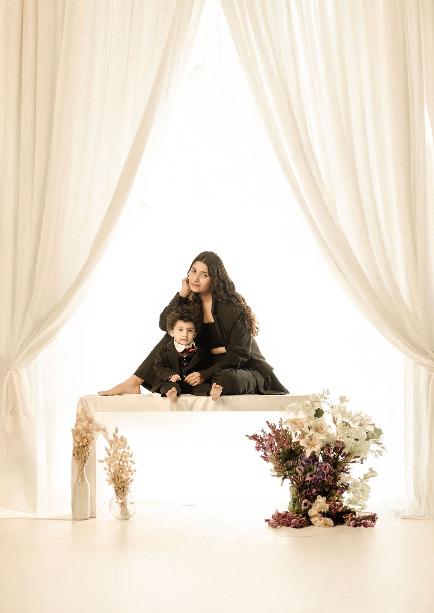 Woman and young boy in black outfits pose elegantly at white table with floral arrangements and flowing curtains backdrop.