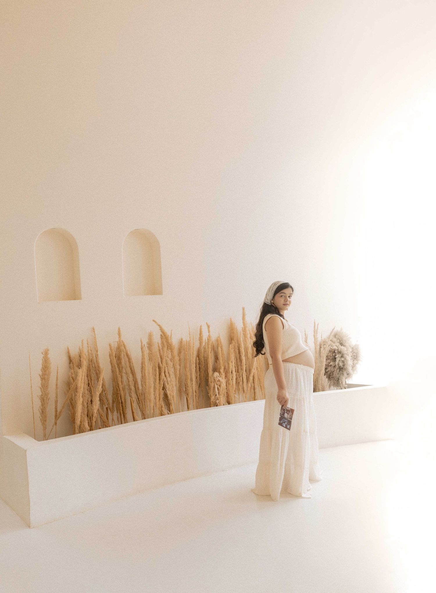 Woman in white crop top and maxi skirt poses beside pampas grass display in minimalist white room with arched niches.