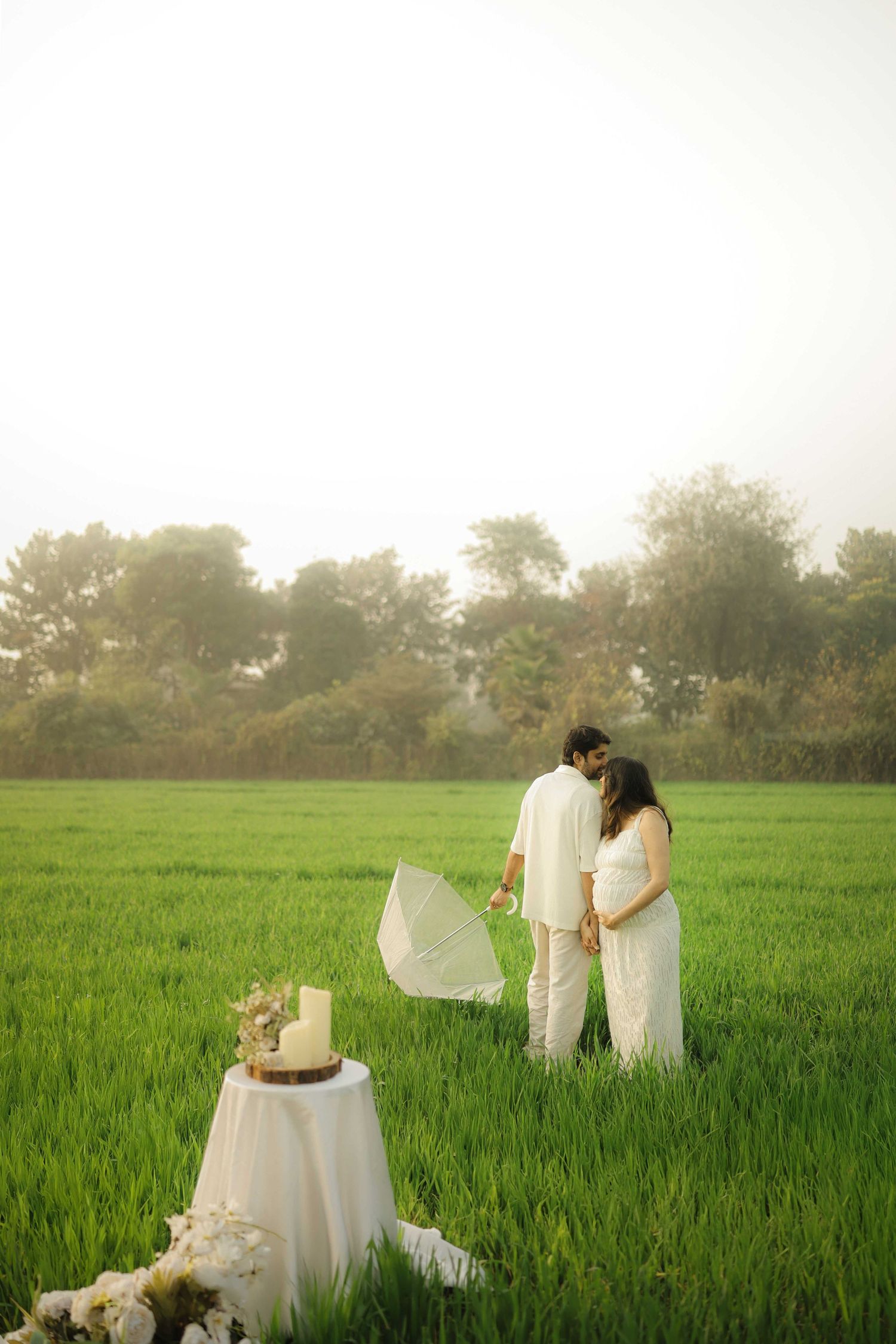 Couple in white attire sharing a tender moment in a lush green field beside a wedding cake on a draped table.