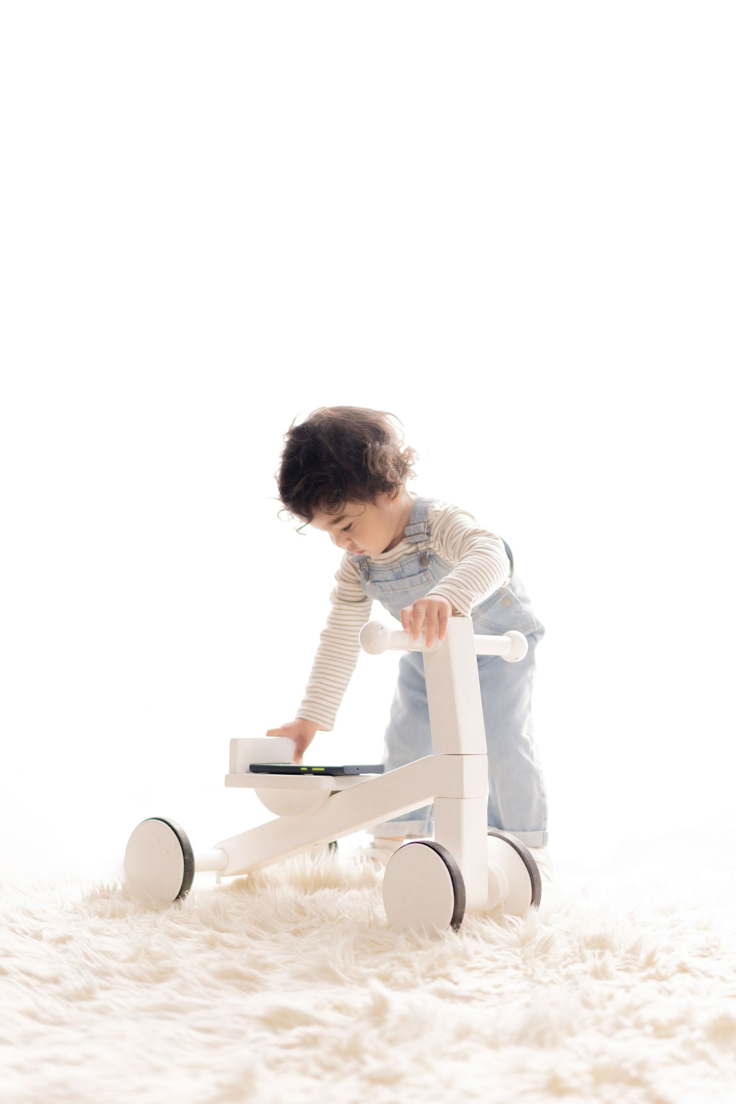 Young toddler in striped shirt and overalls plays with a wooden balance bike on a fluffy white rug.