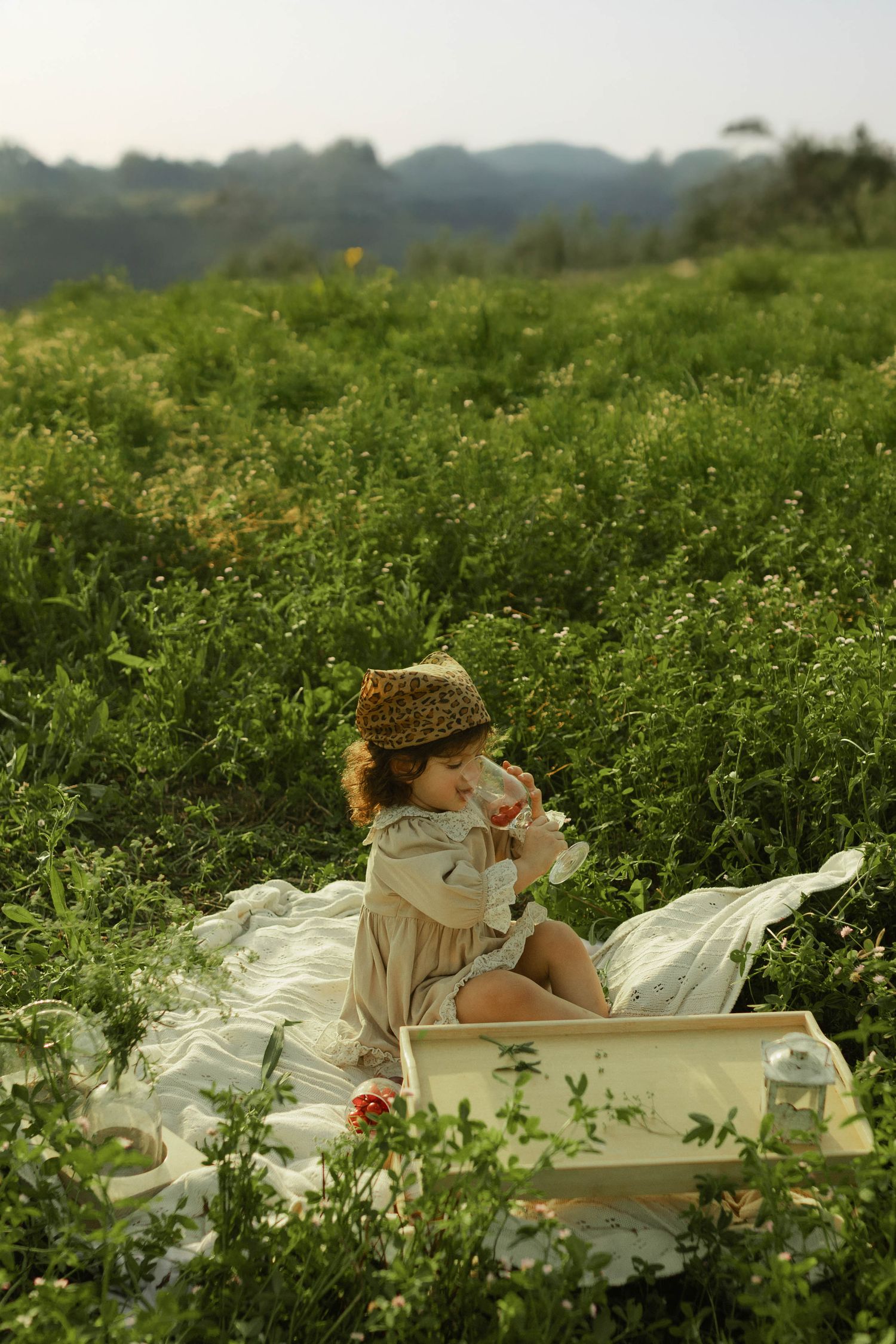 Young child in leopard hat sits on white blanket in lush green meadow, holding a flower during outdoor picnic.