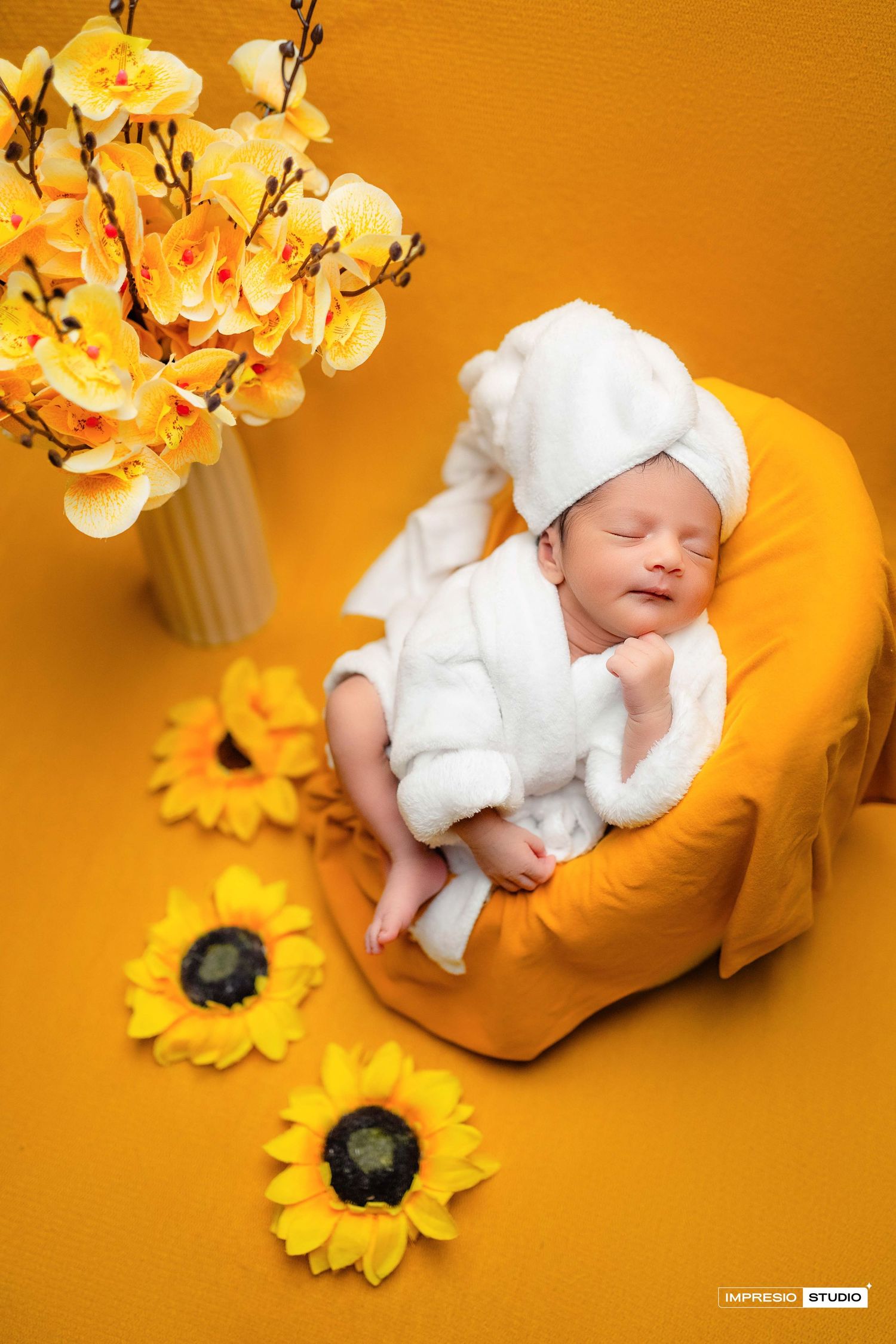 Sleeping newborn baby in white robe and towel turban on yellow bean bag surrounded by sunflowers.