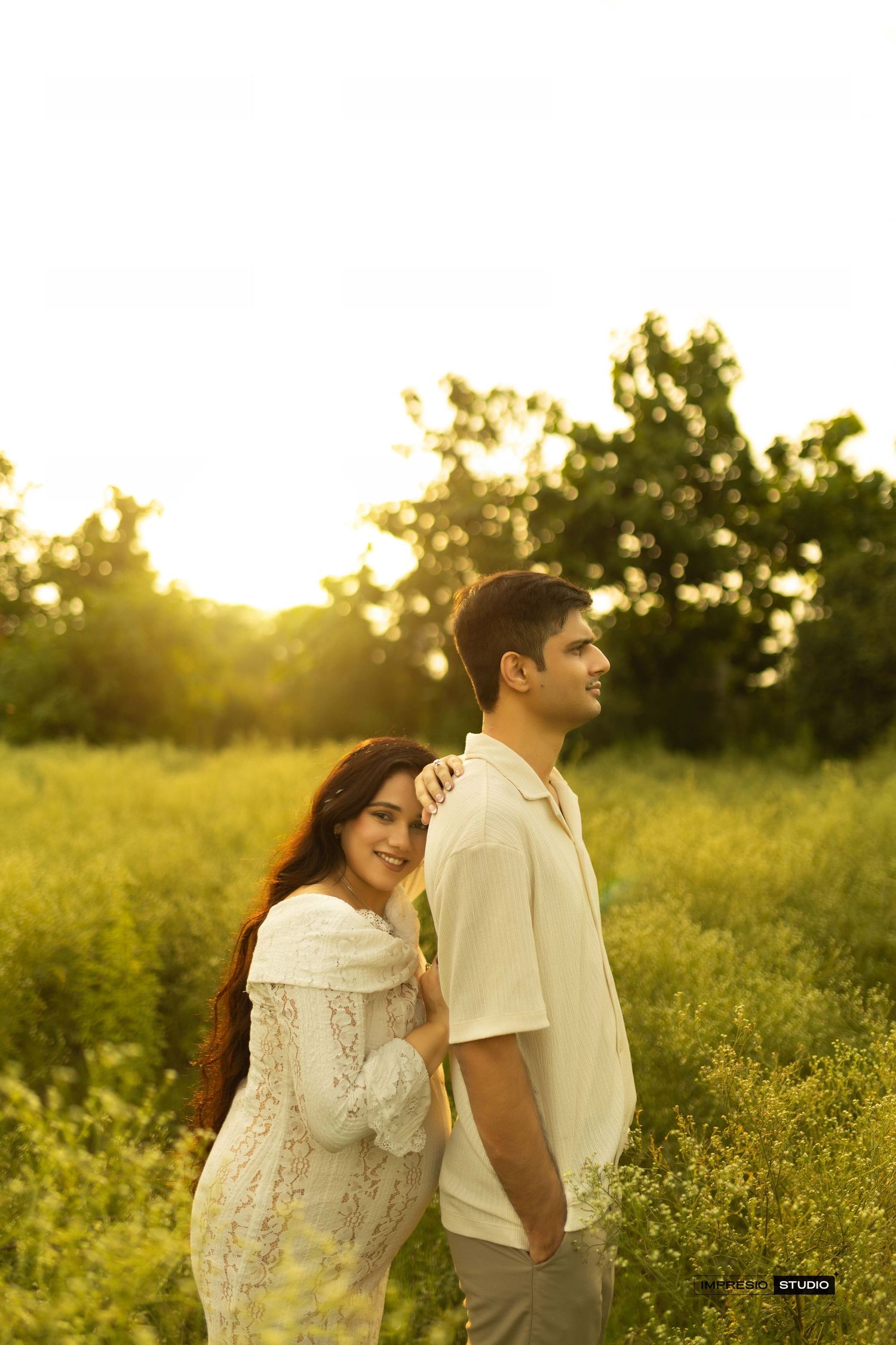 Couple posing in golden hour sunlight in a lush green field, woman in white lace dress smiling, man in cream shirt.