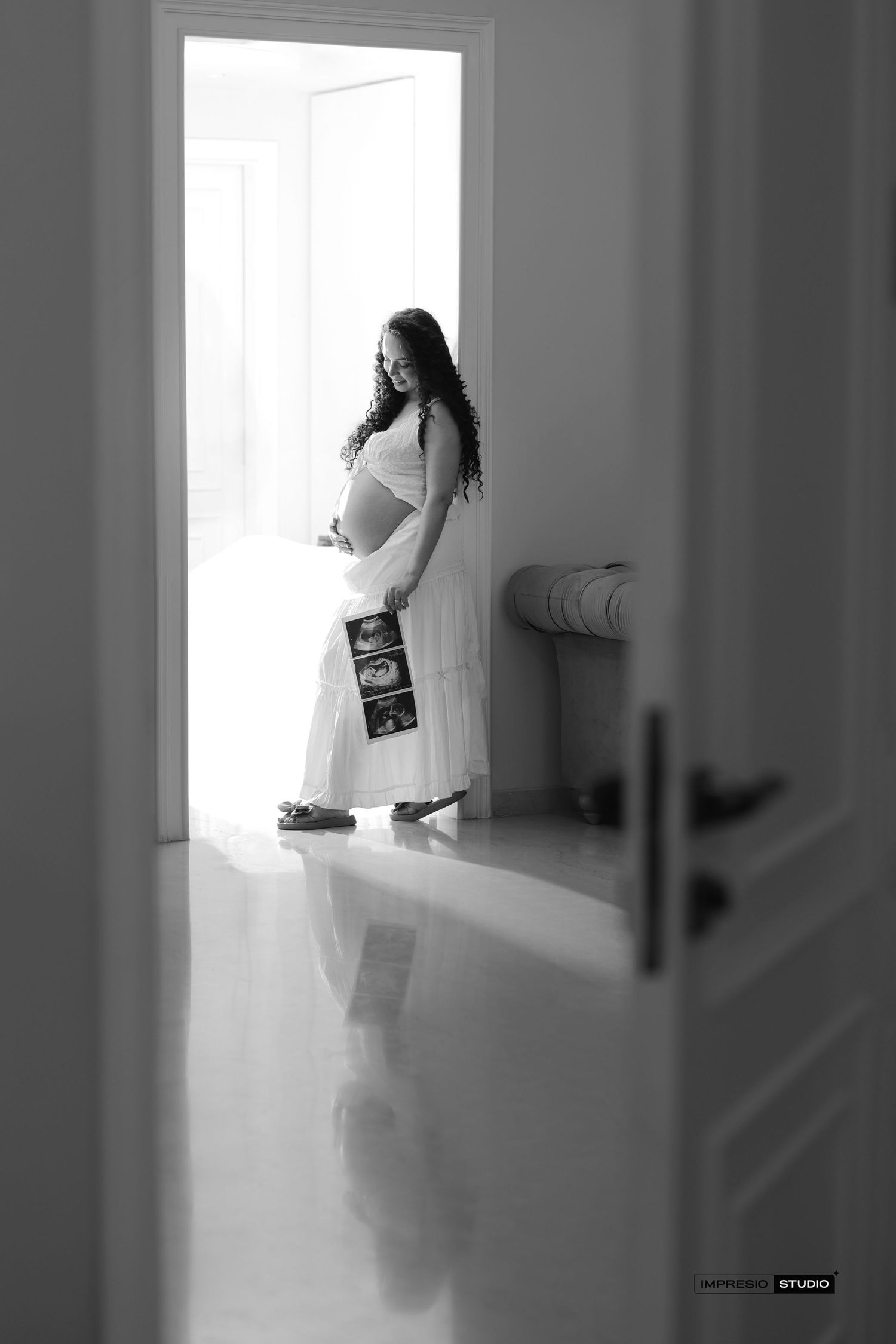 Black and white maternity photo of pregnant woman in white dress holding ultrasound photos, standing in bright doorway.