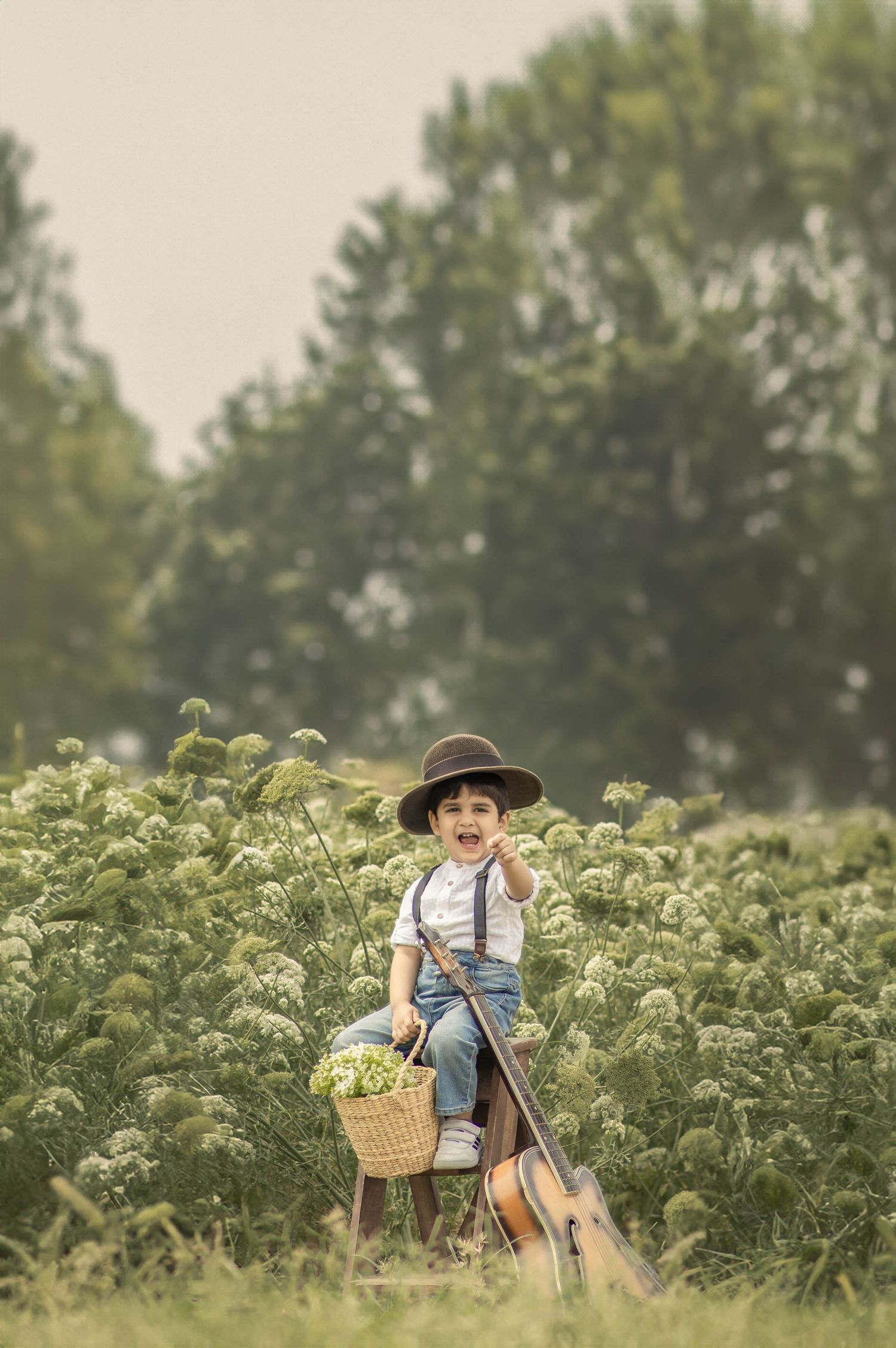 Young boy in hat and suspenders sits on stool in field with flower basket and guitar, smiling outdoors.