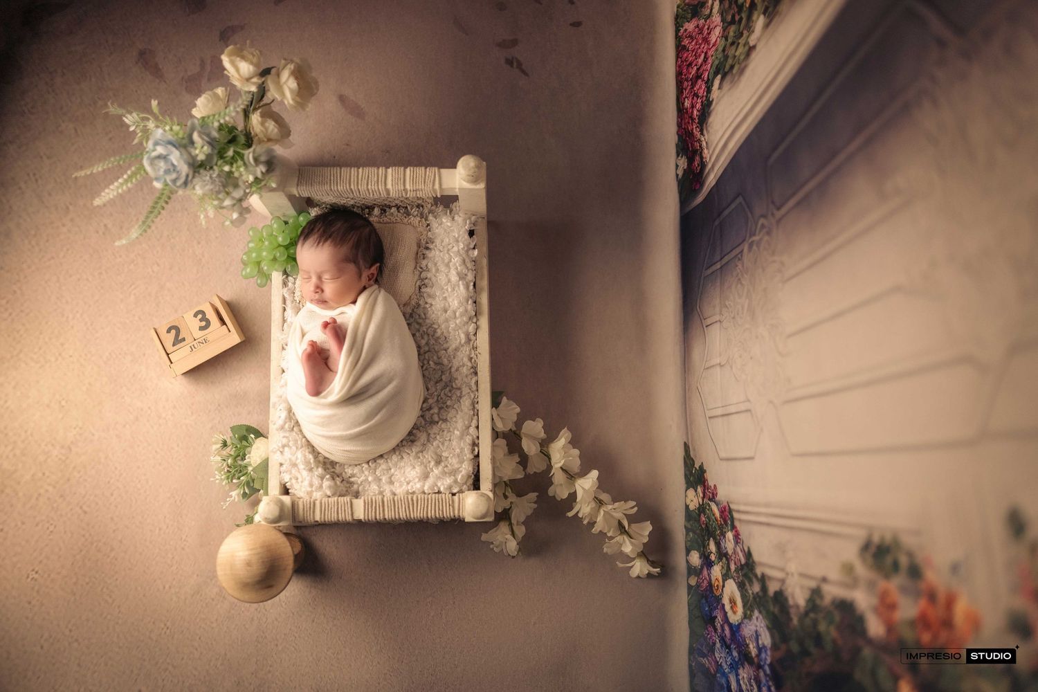Newborn baby wrapped in white swaddle sleeping peacefully in a tiny decorative white crib with floral accents.
