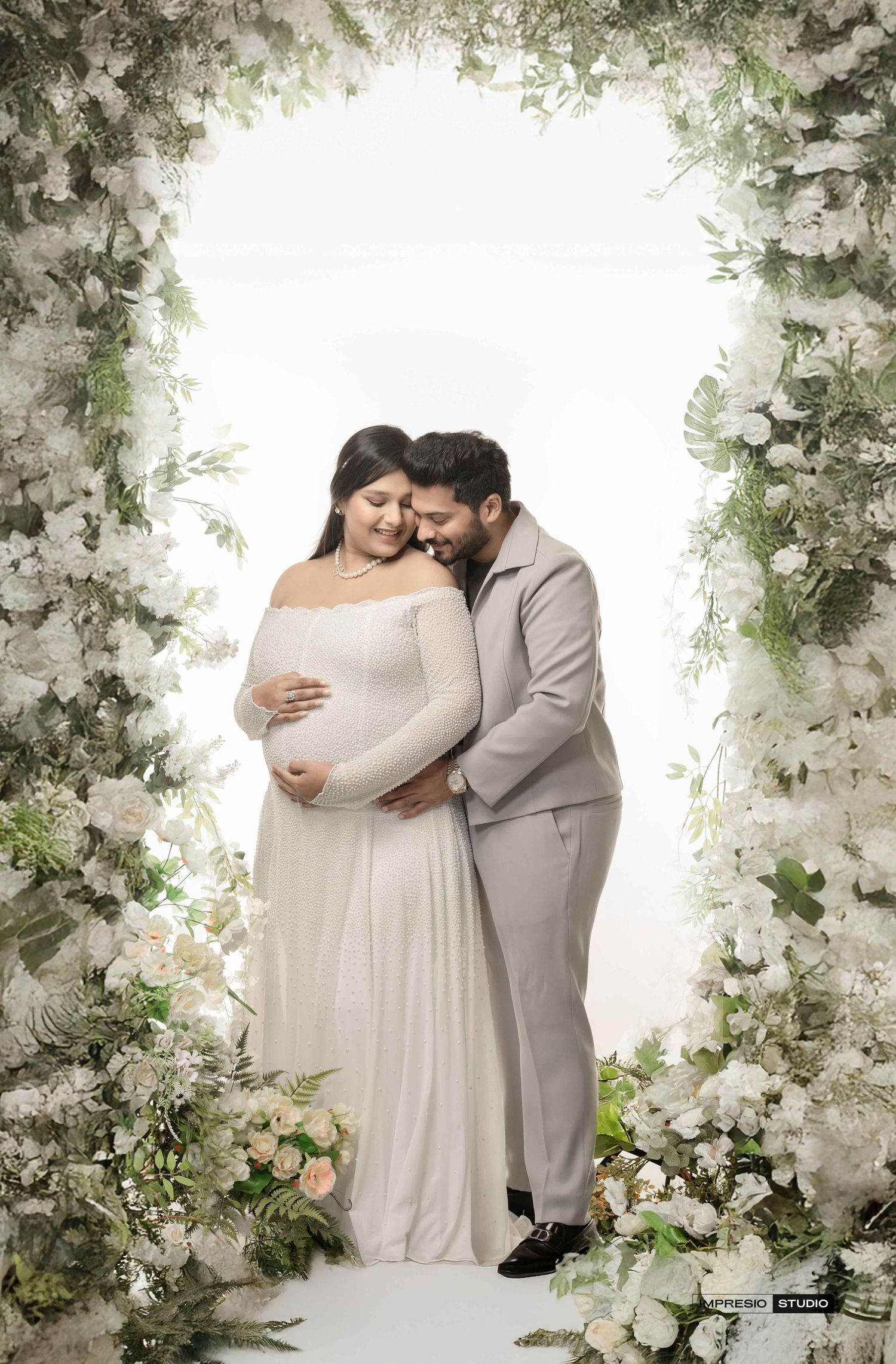 Couple maternity photoshoot with floral arch, pregnant woman in white gown, man in beige suit, studio setting.