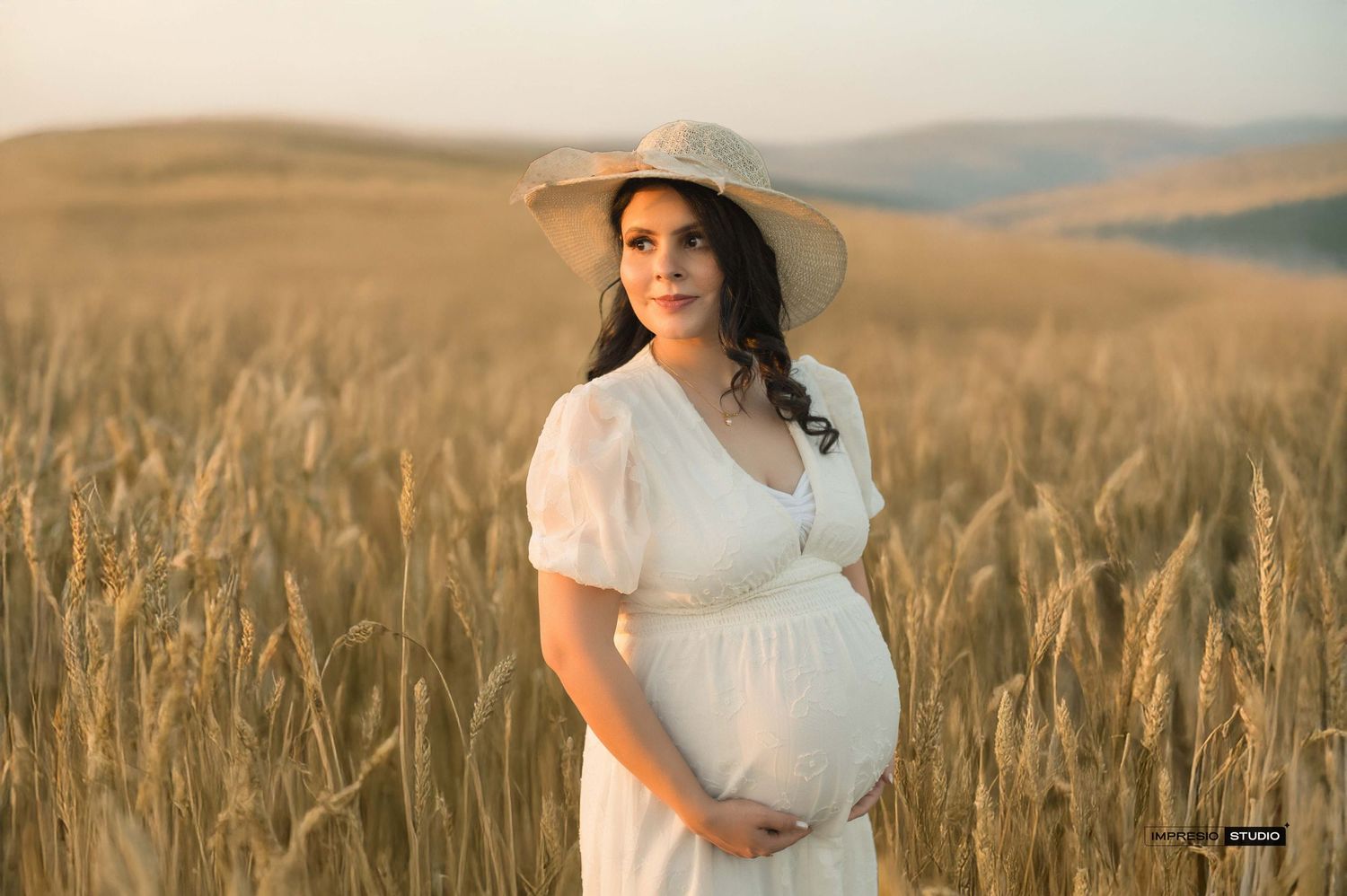 Pregnant woman in white dress and straw hat standing in golden wheat field at sunset, cradling her baby bump.