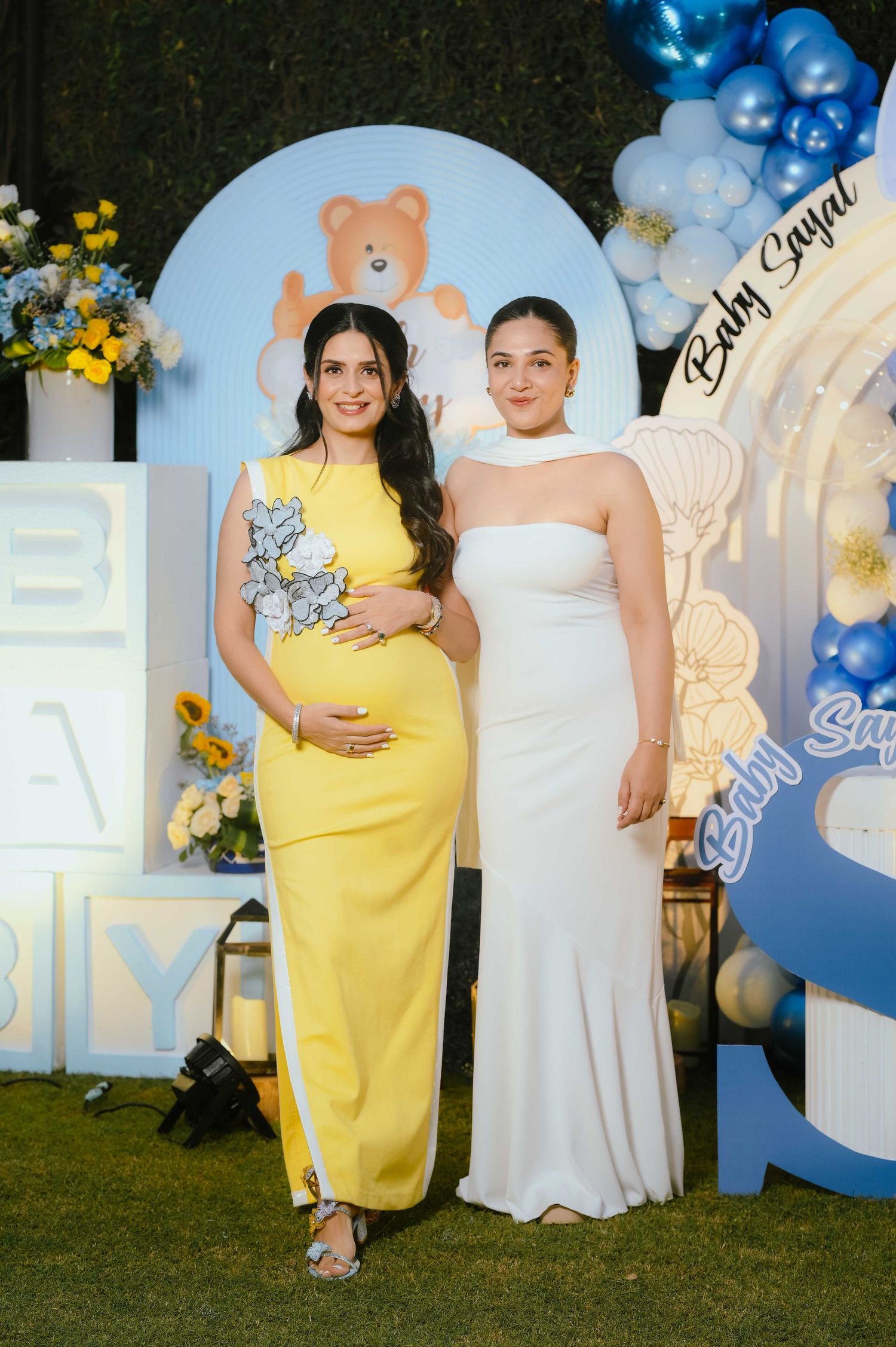 Two women pose at a baby shower, one in yellow maternity dress, beside blue balloon arch and teddy bear decorations.